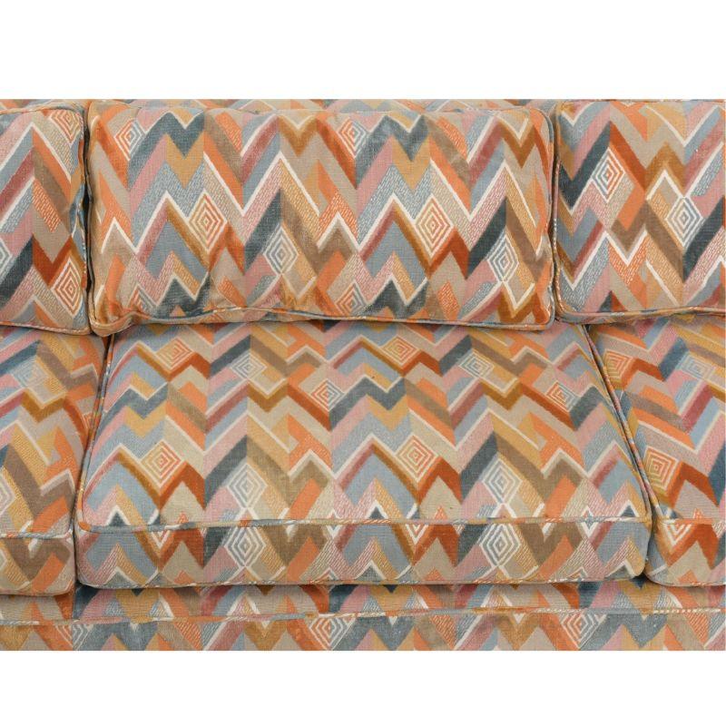 A vintage pair of Dunbar sofas, circa 1970, upholstered in multicolor flame stich chevron fabric.  The distinctive colorful fabric appears to be Jack Lenor Larsen.  The three seat sofas have track arms with a square shape to sides and back.  Each