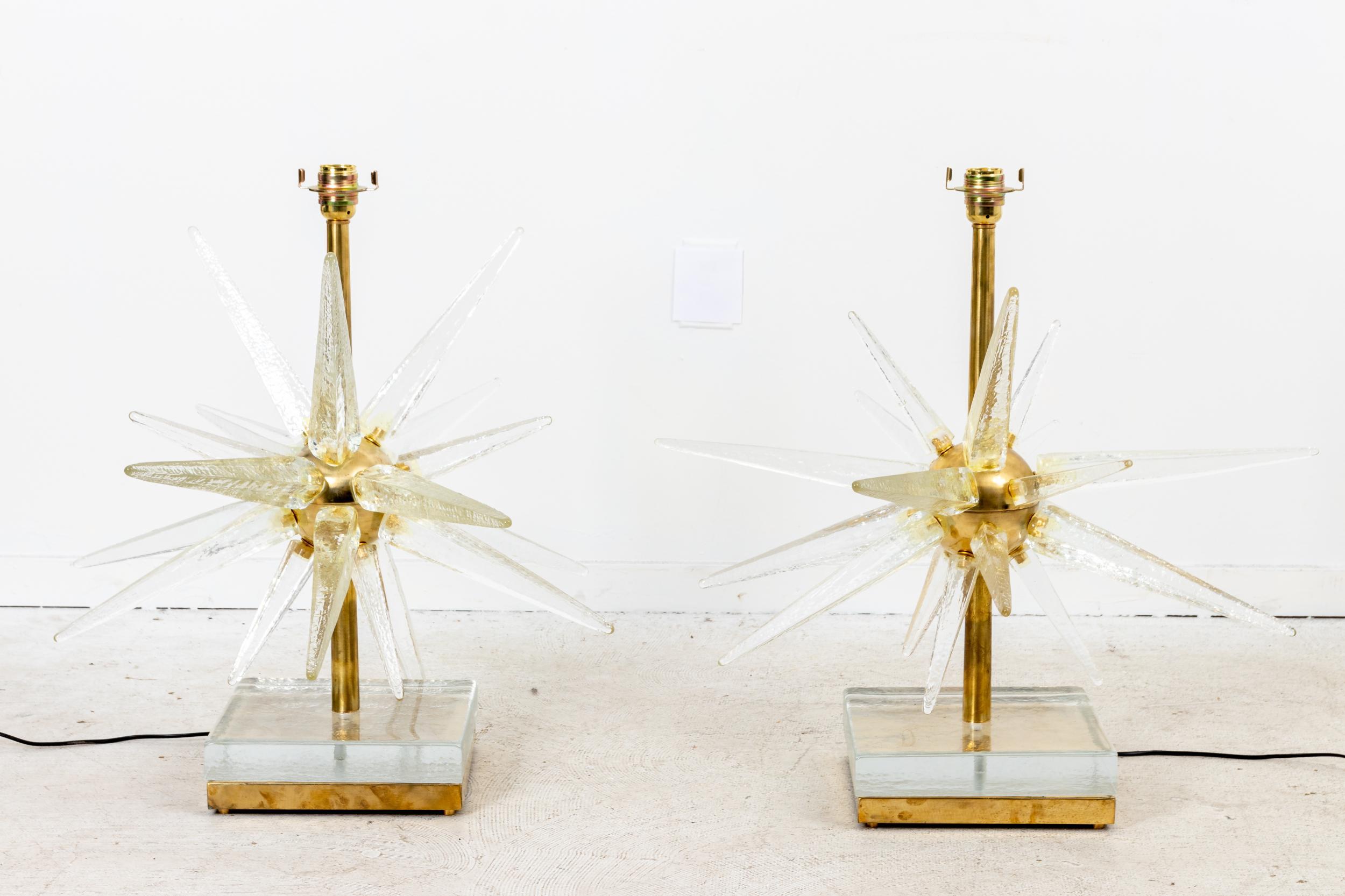 A pair of Murano glass starburst table lamps. This pair is made in Italy with molded glass radiating off a brass center. Each glass ray has slight variations, showing the handmade quality. A thick glass block sits atop the brass base, reflecting a