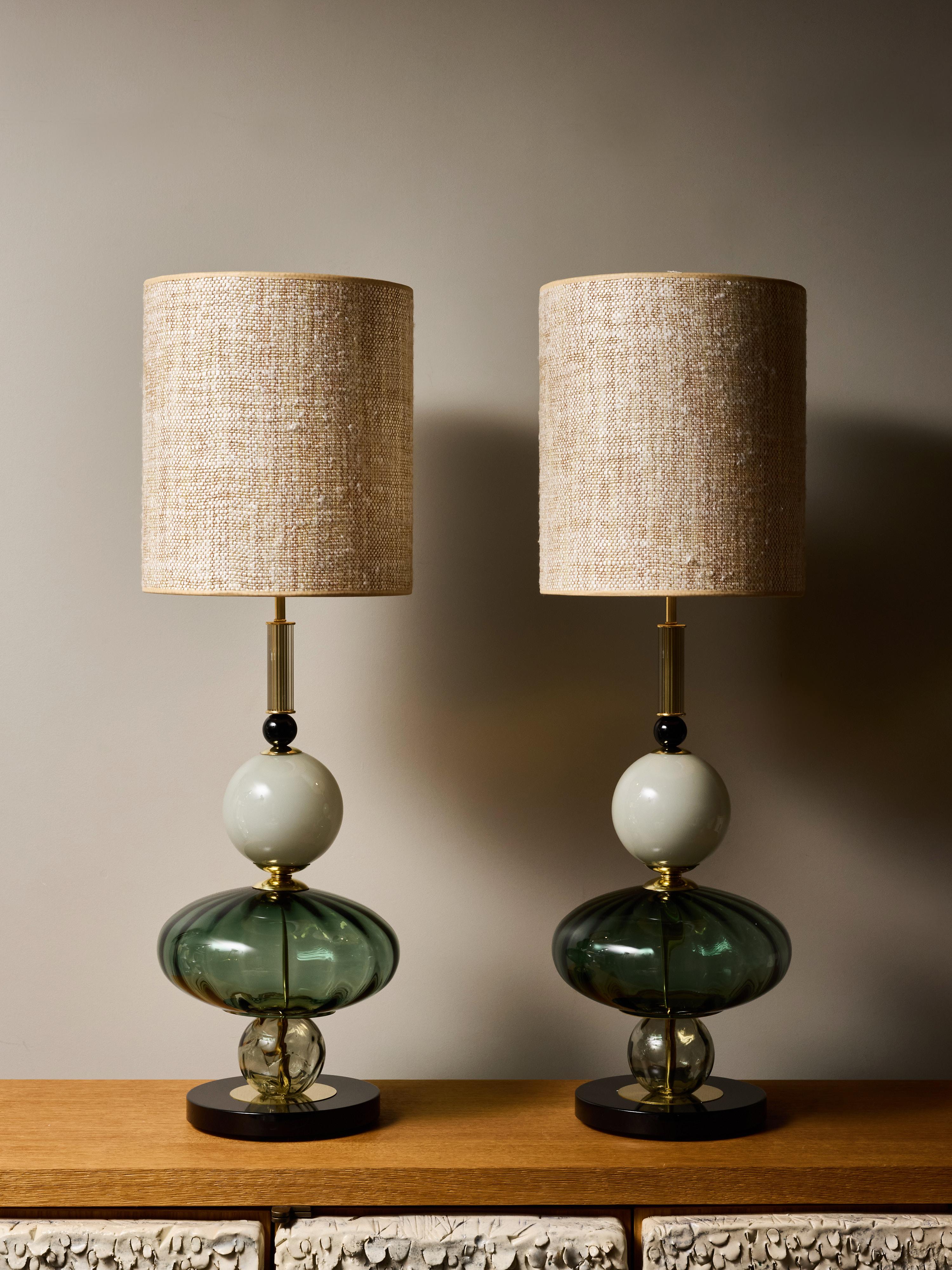Este par de lámparas de sobremesa artísticamente compuestas presenta una sofisticada interacción de color, forma y material. Cada lámpara se construye en torno a una escultural pila de elementos de vidrio soplado a mano: una esfera opalina verde