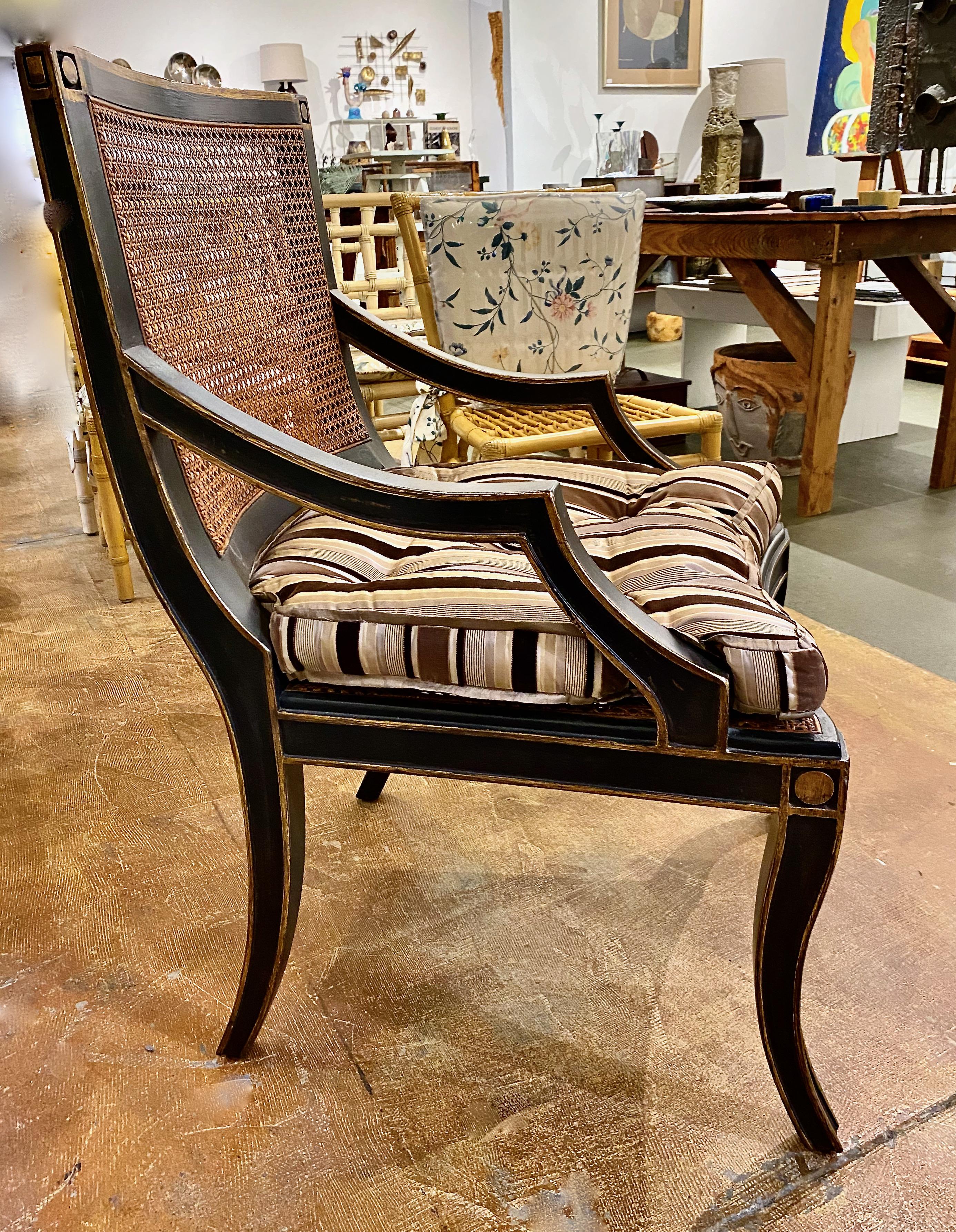 This is an outstanding pair of early 21st century open arm chairs in the Hollywood Regency style. The chairs feature ebonized carved frames with caned seats and backs. The chairs are detailed in gold leaf over the ebonized frames. Both chairs are in
