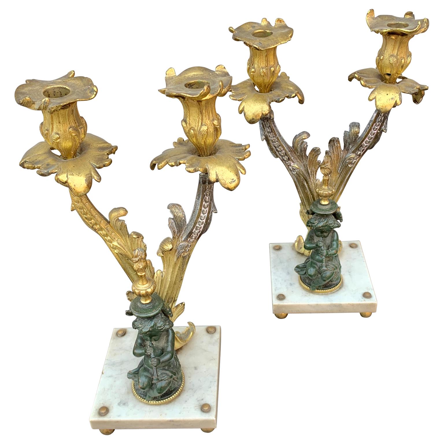 19th century pair of French gilded and bronze two-armed candelabra with Carrara marble square base and cast iron painted putties playing the flute. 
From the second part of 19th century. They are presented in old patina and unpolished to keep the