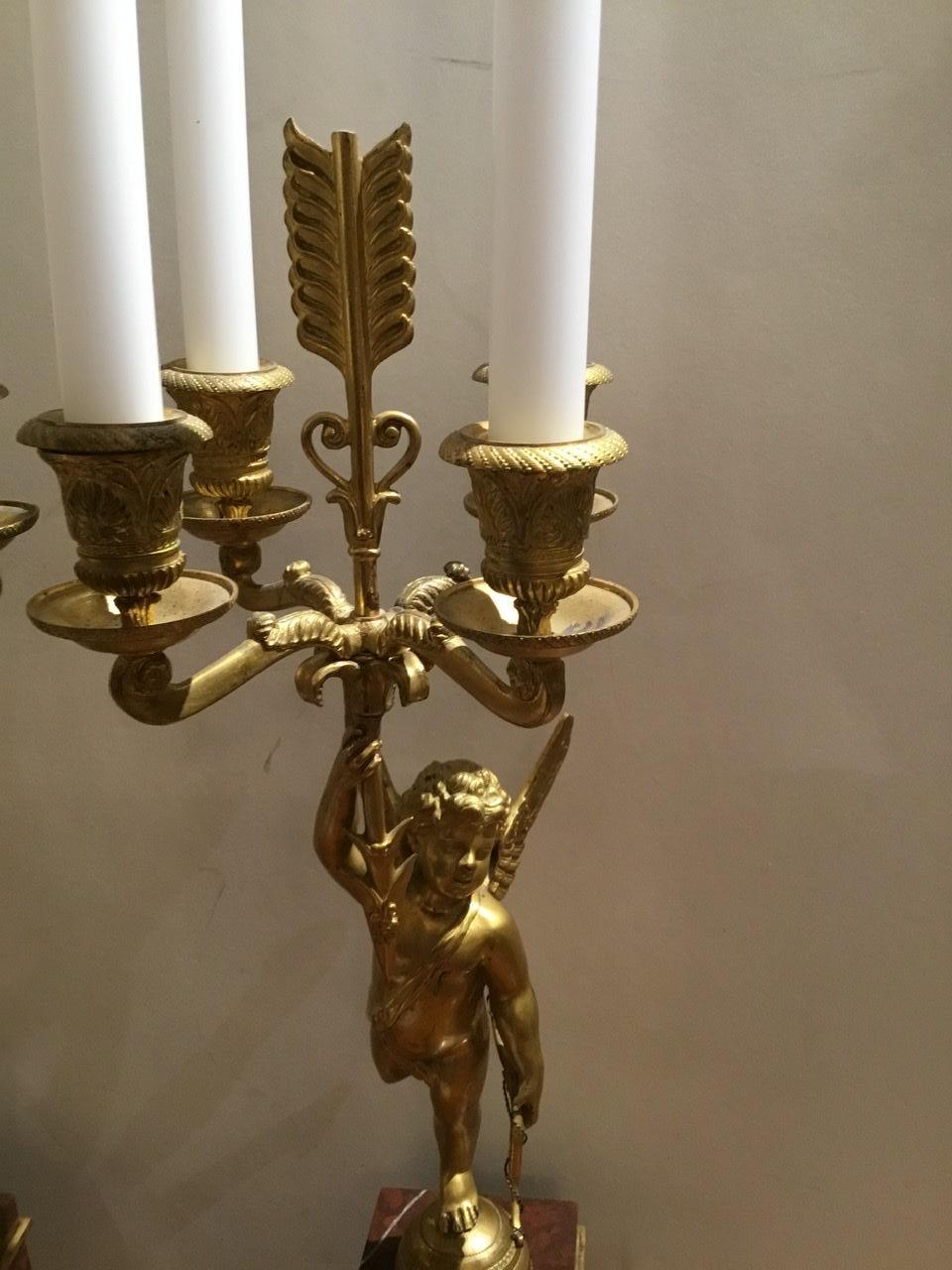 Empire style four light candelabrum, cupid-form standards poised on
Turned half spheres, holding a bow in one hand and an arrow supporting
Candle arms set with anthemion-molded cups in the other, the marble
Bases decorated with bronze appliqués