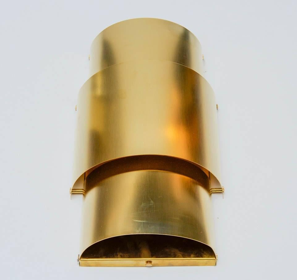 Pair of wall sconces made of natural brass with two inner lights per sconce.

Straight backplate on which are coming two curved brass sheets, with the light escaping from the gap between them.