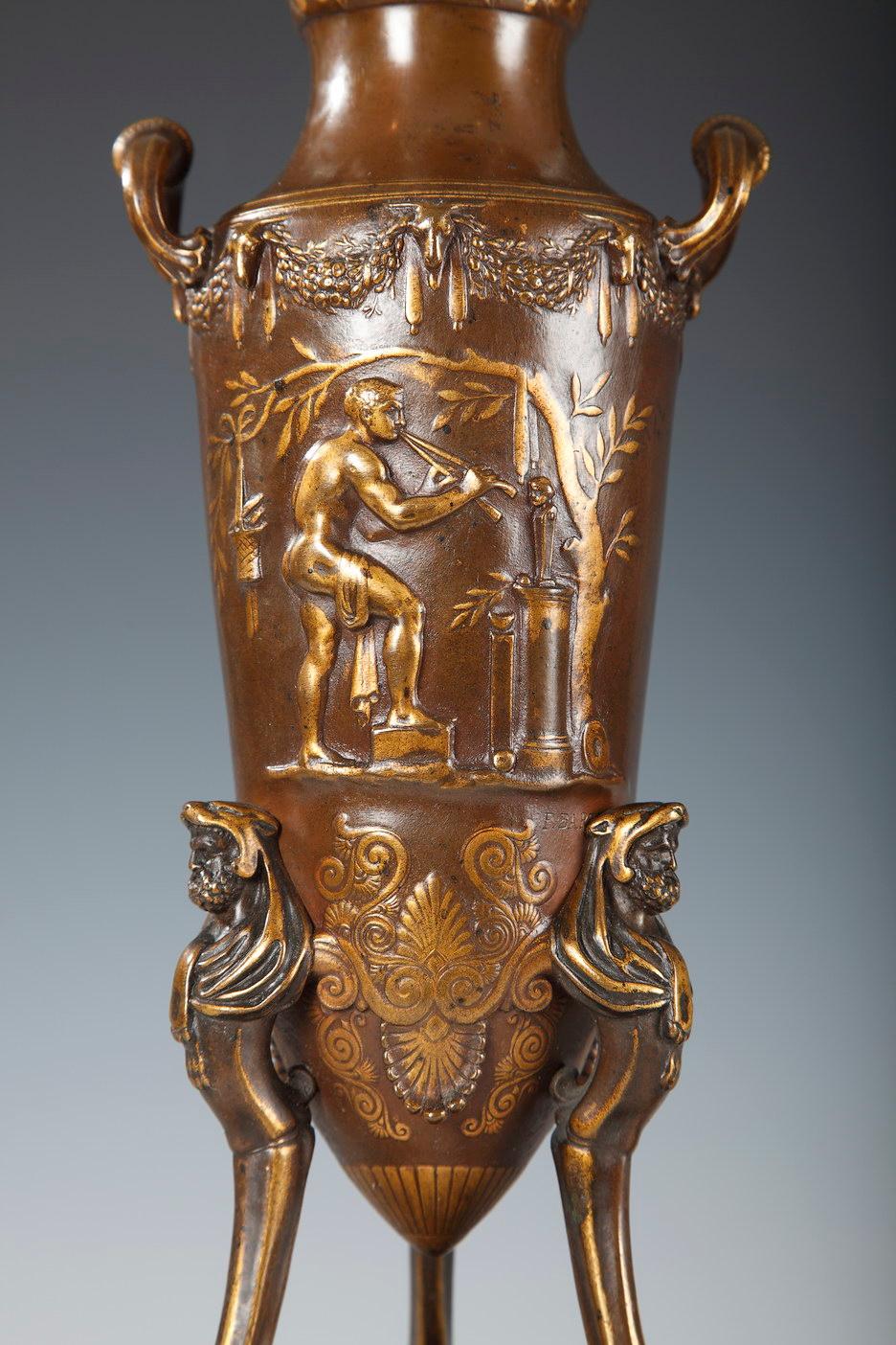 Paire de vases de style grec en bronze à deux patines. Chacune, conçue en forme d'amphore tripode ornée de têtes d'Hercule, est décorée en bas-relief de scènes de sacrifice de l'Antiquité. De nombreux masques, pampres, instruments de musique et