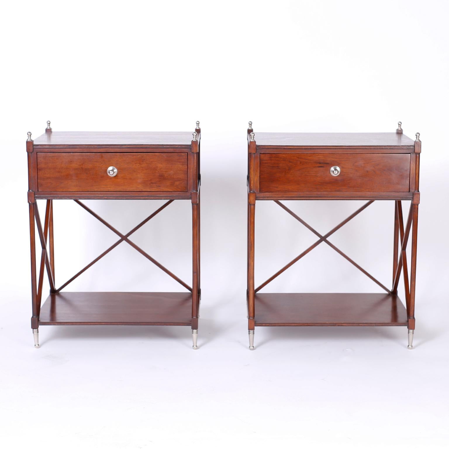 Pair of neoclassical style one-drawer stands crafted in mahogany with side and back X-supports, lower storage tier, and silvered metal finials and feet.