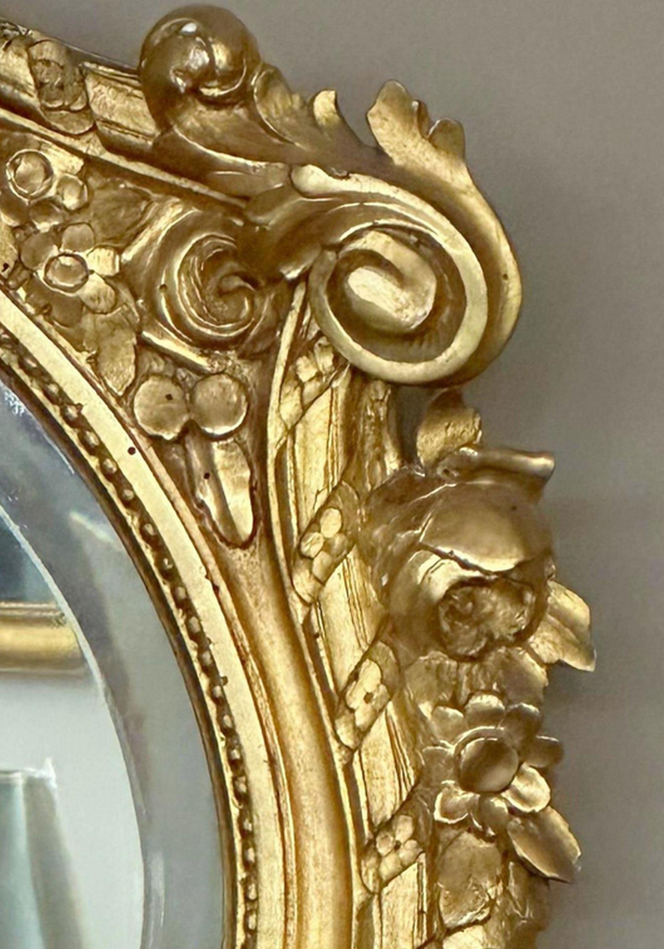 Fine pair of 24-karat giltwood mirrors. The oval beveled plates within molded and cabochon borders and surmounted by a cartouche and floral foliate cresting.