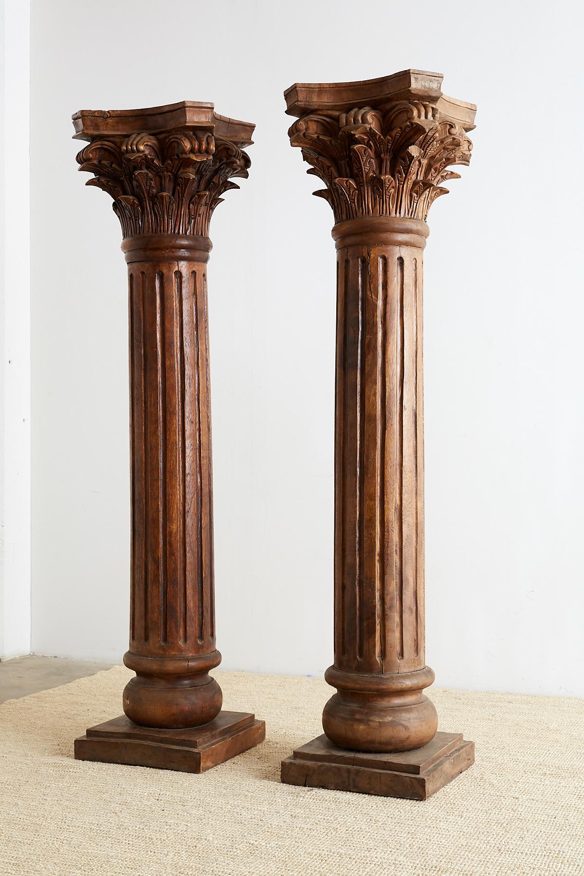 Anglo-Indian pair of hand-carved solid teak columns made in the neoclassical Greco-Roman style with Corinthian capitals. The Corinthian order capitals are supported by large, fluted columns that rest on a square base. The tops showcase large
