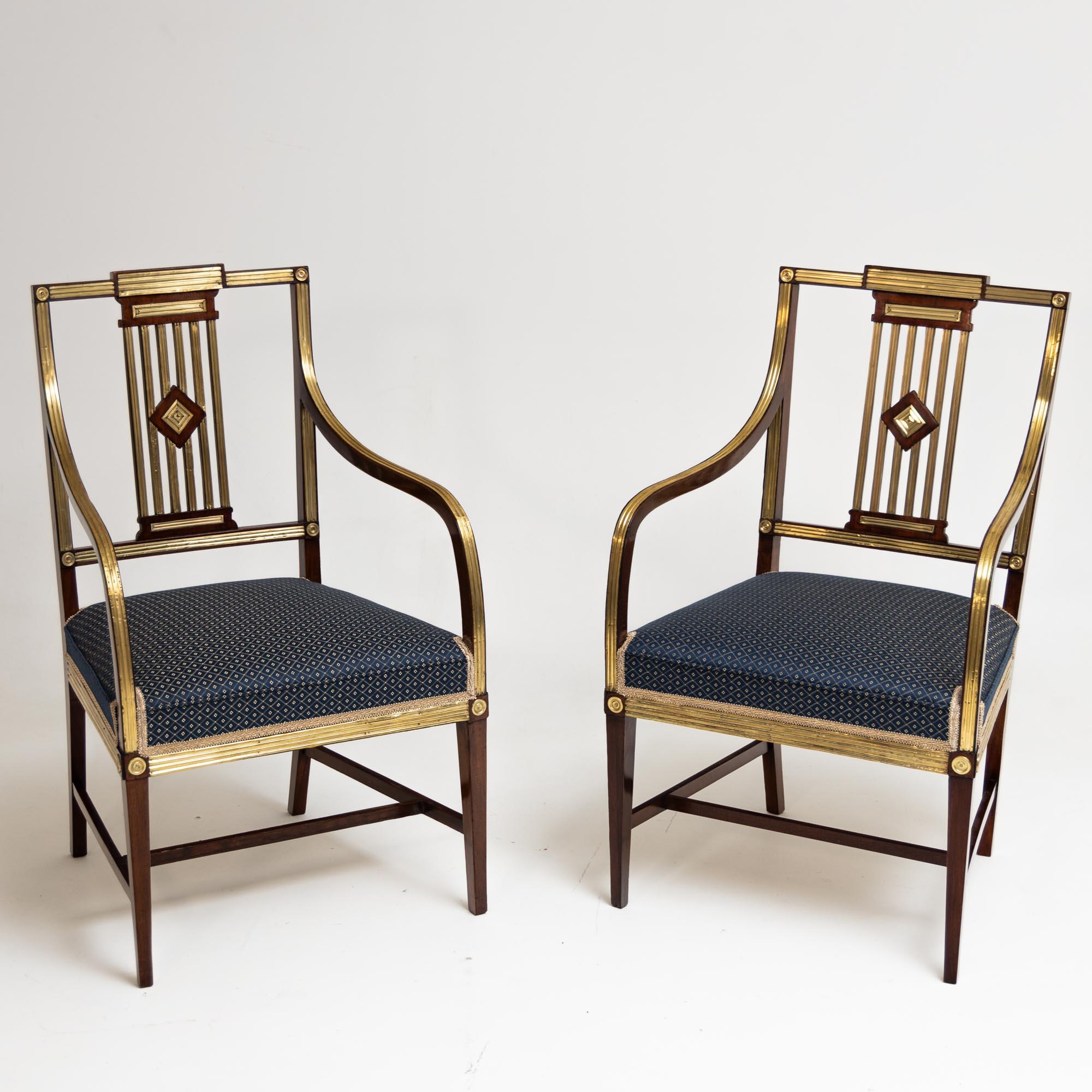 Two classicistic mahogany armchairs with brass decoration and openwork backs. The chairs stand on square pointed legs with H-shaped bracing, which merge into elegantly curved, high-set armrests with gilded decoration. The seats are reupholstered and