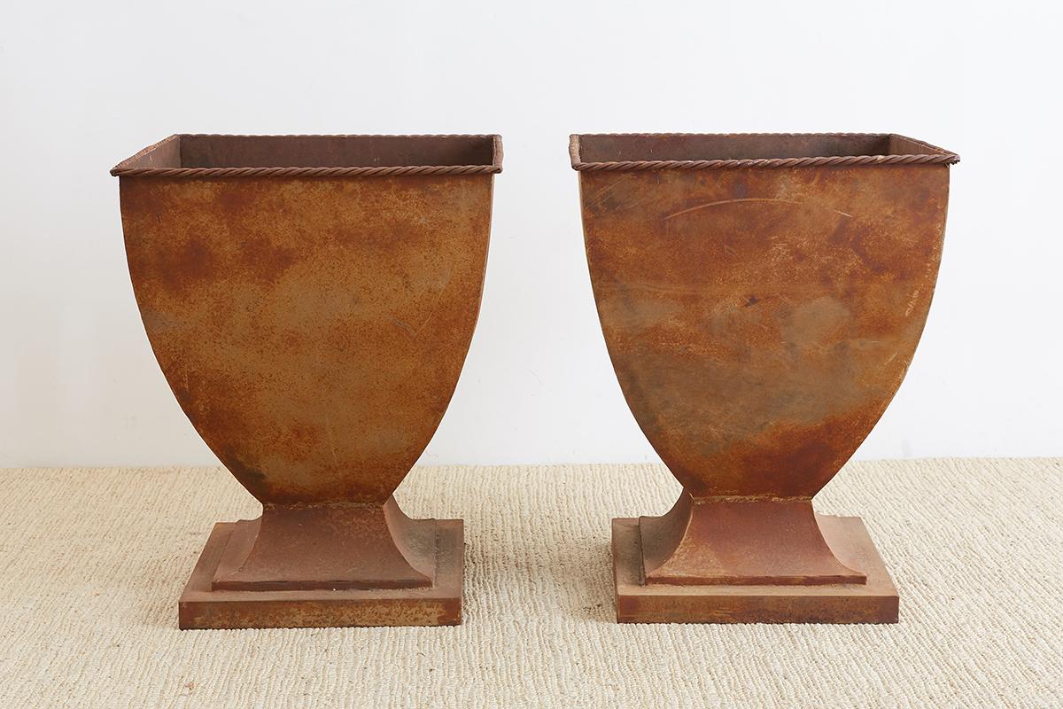 Monumental pair of iron variform garden urns, planters, or jardinière’s. Featuring a tapered square body with ad decorative faux rope design on the top lip. Made in the Greco-Roman neoclassical taste with lovely patinated metal patina. The inside