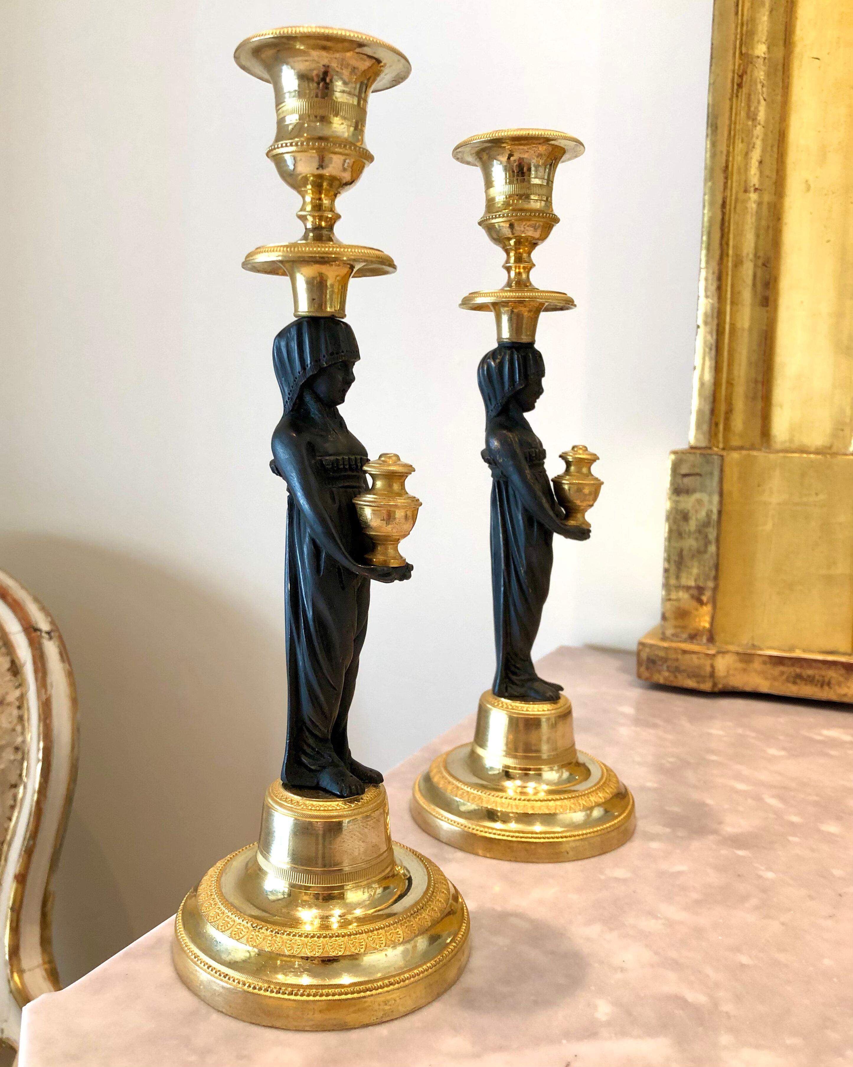 A very rare and excellent pair of ormolu and patinated bronze candlesticks, with the representation of a caryatid vestal virgin wearing a classic tunic and holding golden oil jars between the arms.
In ancient Rome, the Vestal Virgins were