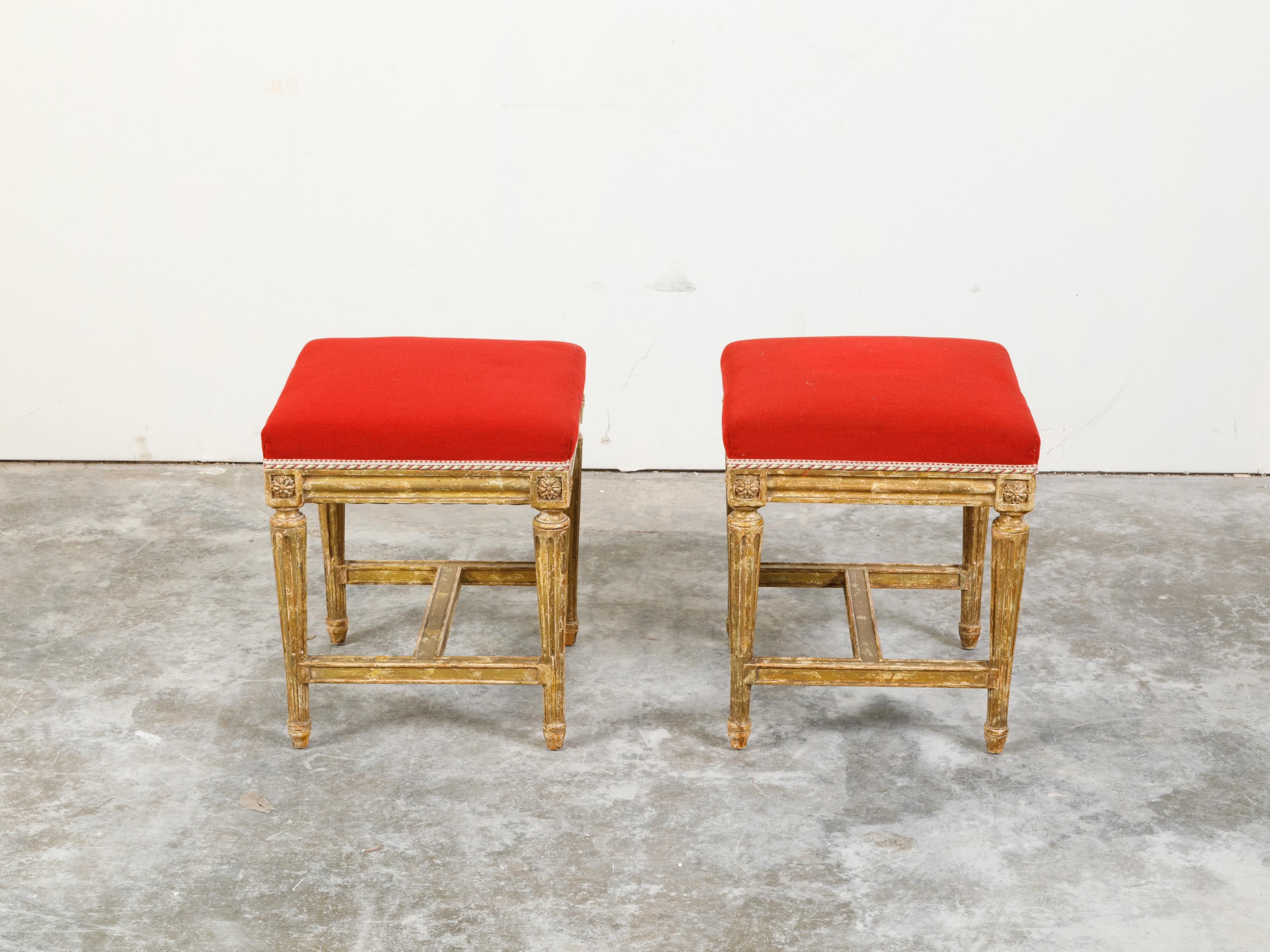 A pair of Neoclassical style painted wood stools from the 19th century, with fluted legs, carved rosettes and red upholstery. Created during the 19th century, each of this pair of Neoclassical style stools features a square seat upholstered with a