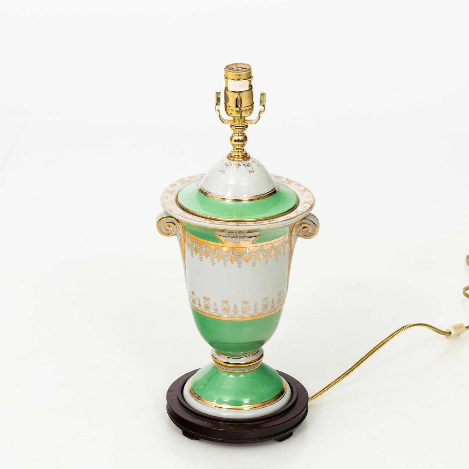 Pair of 1930s green white and gold neoclassical style ceramic lamps. Urn form on wooden bases. There is one minor old repair on one of the handles. Shades not included.
 