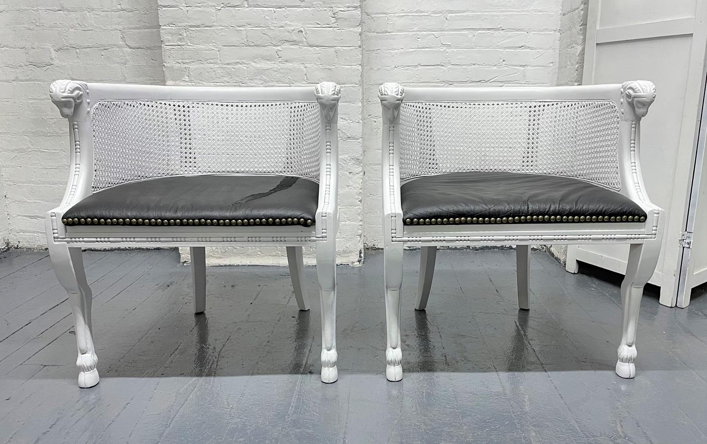 Pair of Neoclassical style ram's head chairs with black vinyl seats. The chairs have caned backs and sides with a solid wood, white painted frame. Carved ram's head to the front of the arms.