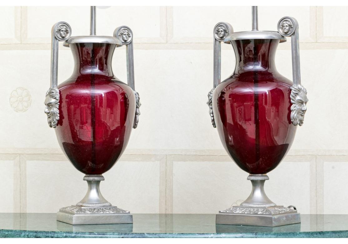 A very stylish pair of Neoclassical style lamps with nickel plated brass mounts over urn form deep ruby red glass vessels. The lamps with two emitting arms with on/off switches. The lamps with dual handles having well cast neo-classical images at