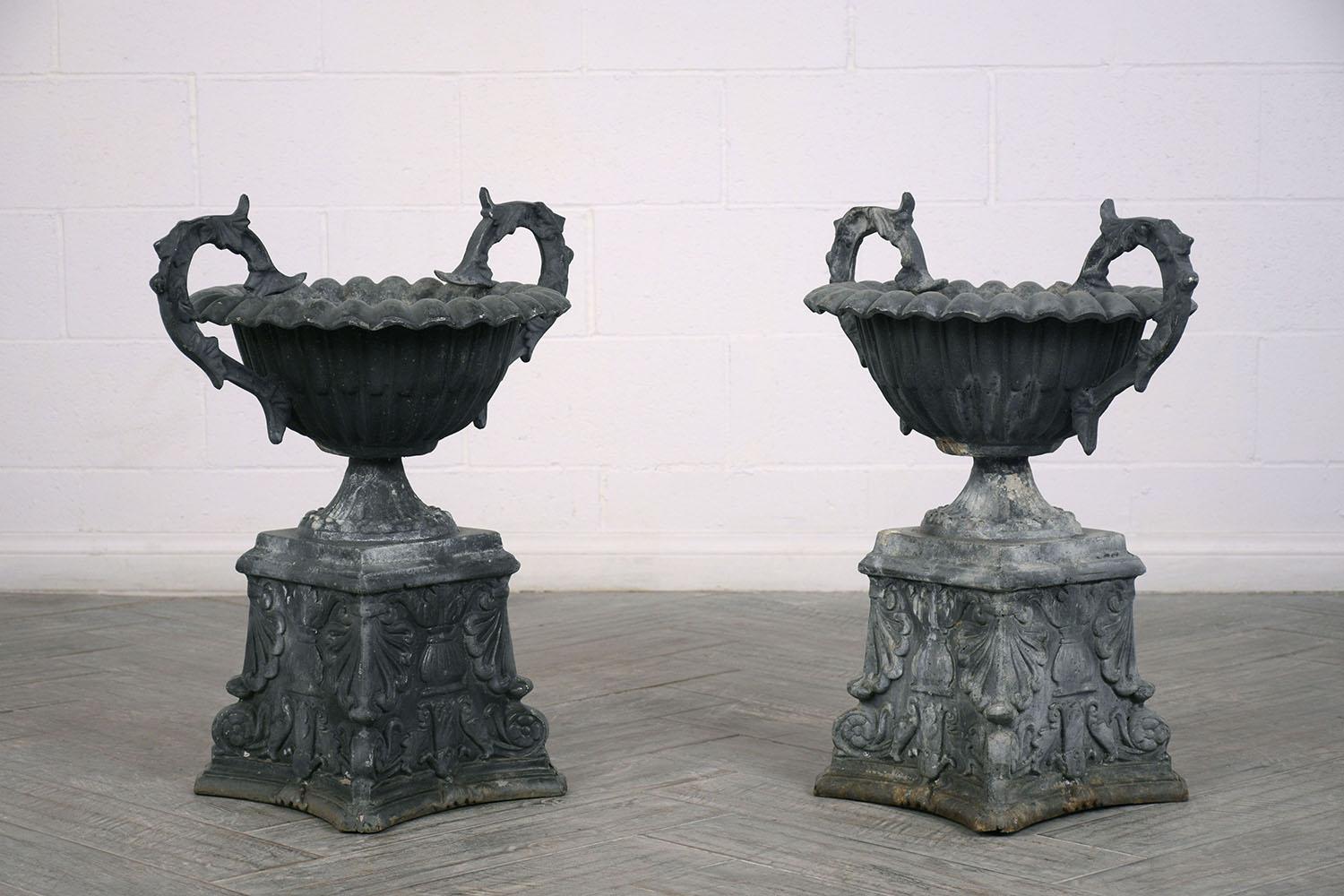 This pair of 1930s neoclassical style outdoor planters or patio garden urns are made of metal with a natural oxidized patina. The urns have two handles with acanthus leaf details and a scalloped lip on the basin. The pedestal is adorned with sea