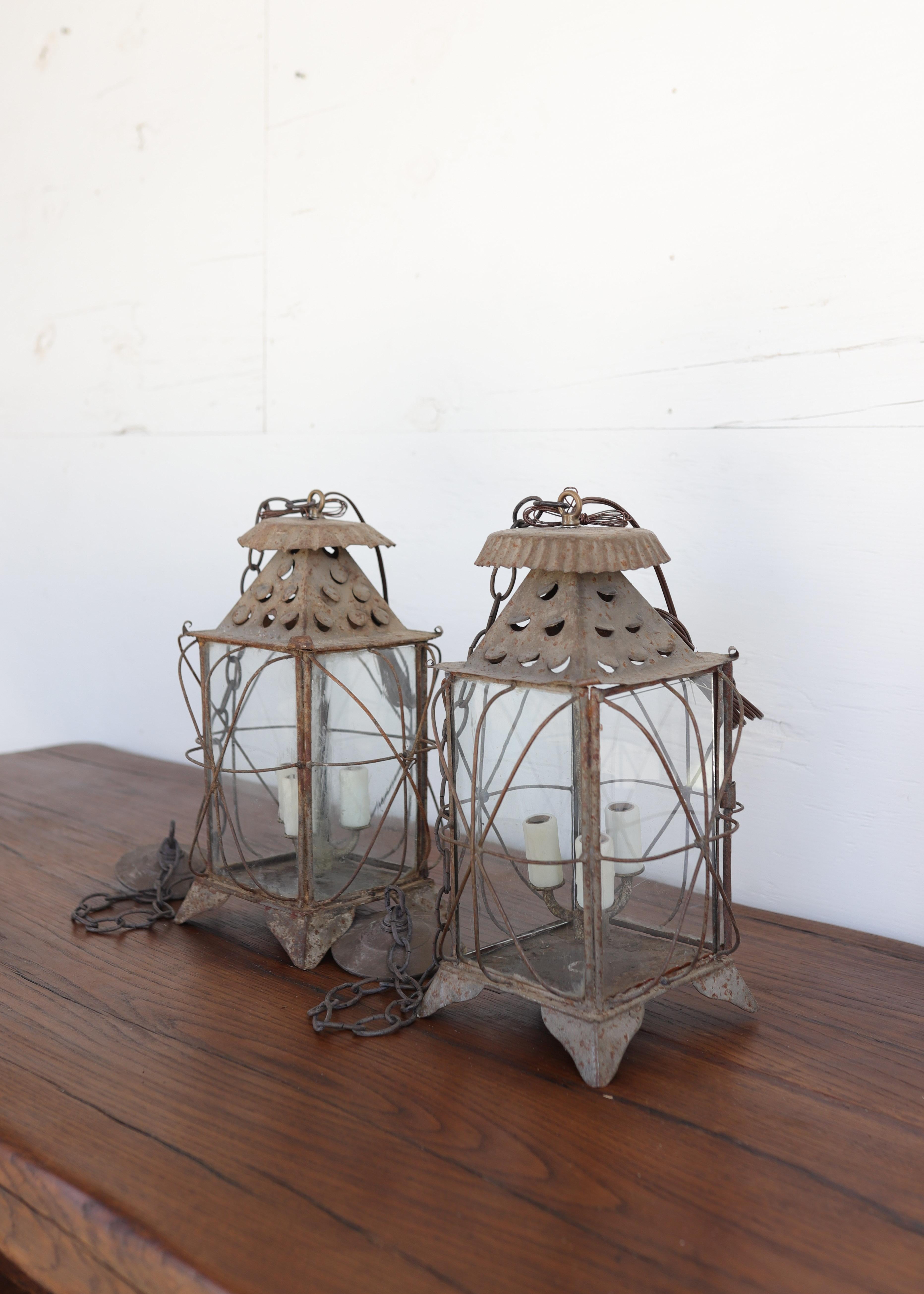 A charming pair of antique lanterns from the South of France, crafted in hand-riveted tôle with original glass panels and a beautifully aged patina. Each lantern features an elegant quatrefoil wire cage design surrounding the tapered body, topped