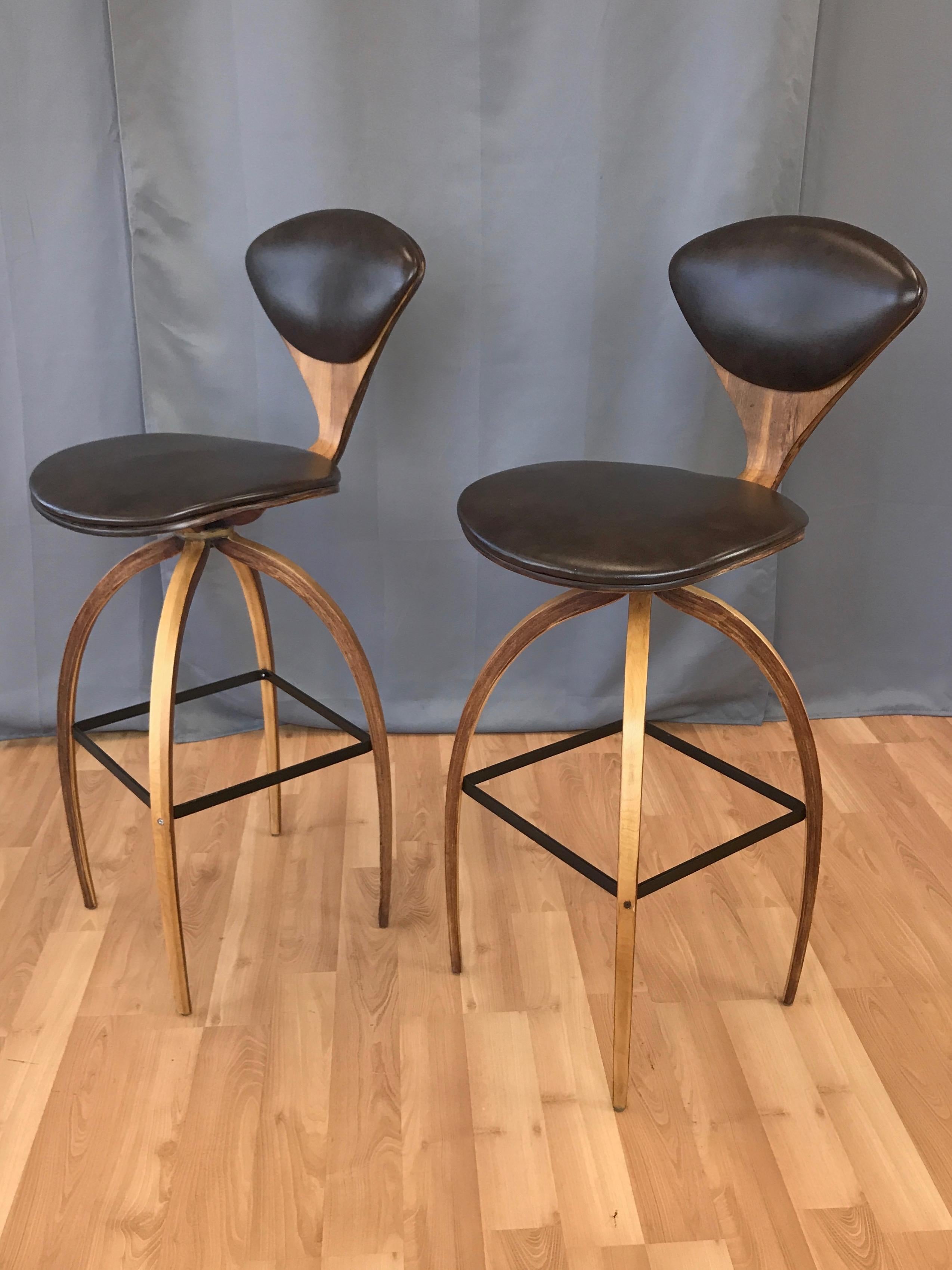 A vintage pair of rare bar height swiveling stools by Norman Cherner for Plycraft.

Comfortable bentwood seat with incredibly slender waist in walnut veneer and mottled dark brown faux leather upholstery. Lithe, two-tone, bentwood spider legs