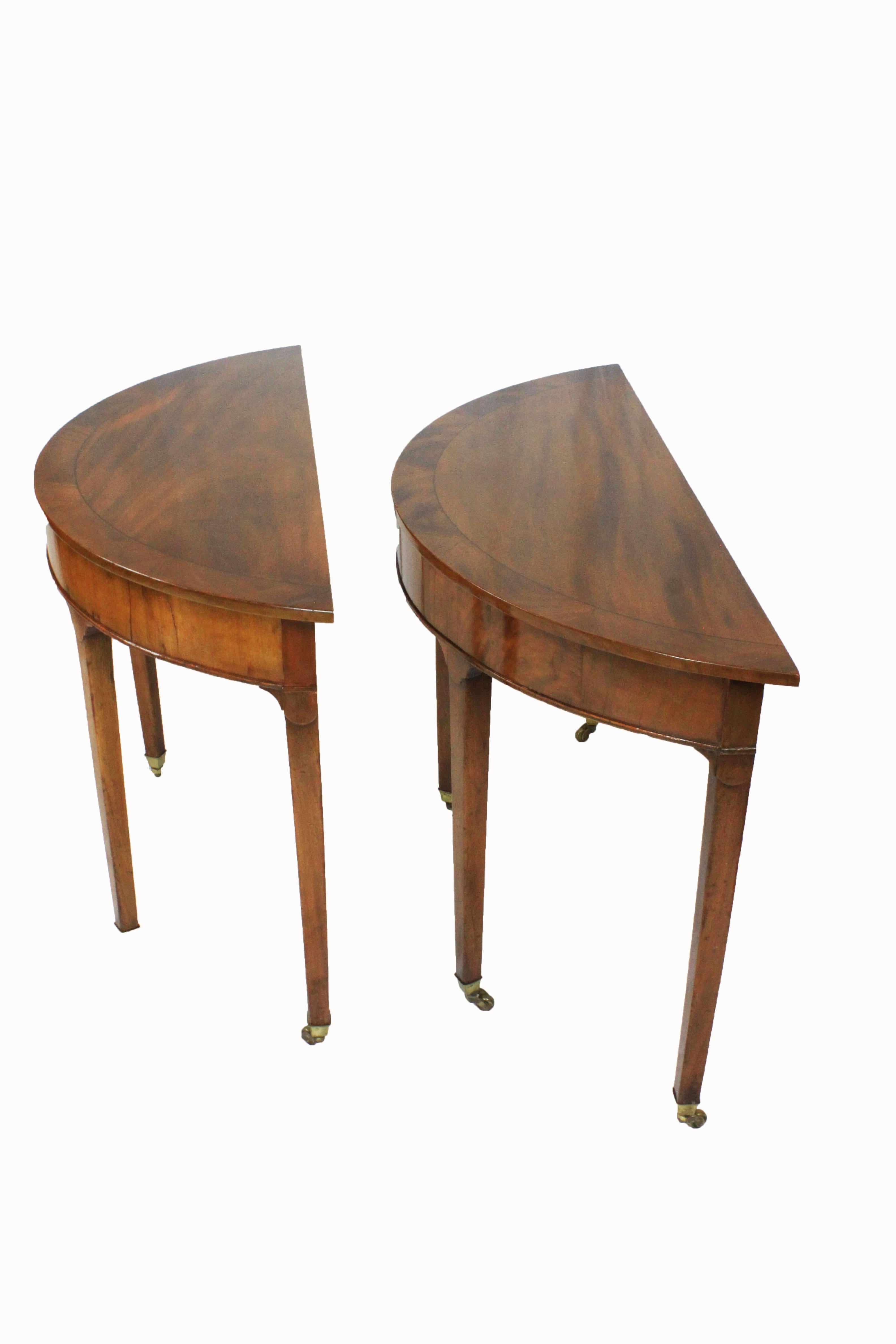 Matching pair of George III mahogany demilunes/consoles crossbanded in mahogany and standing on slender tapered legs terminating with brass castors. Warm patinated plum pudding mahogany tops with mahogany crossbanding.