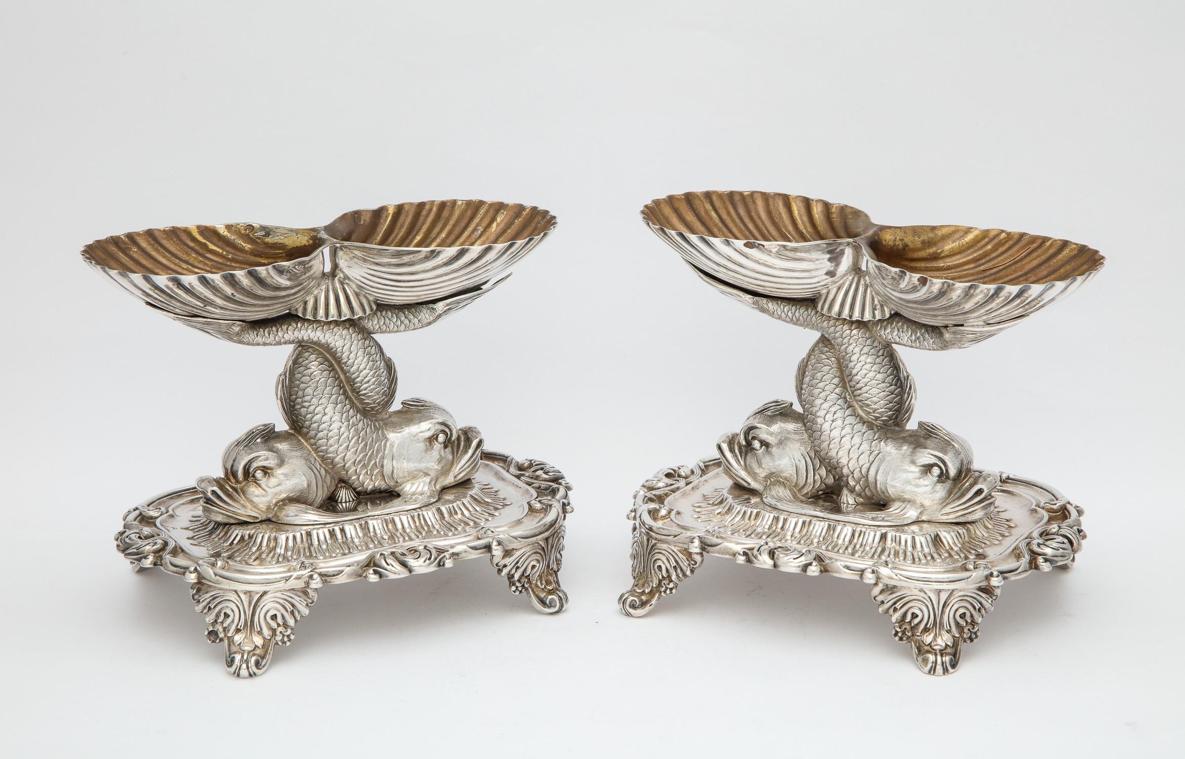 Pair of sterling silver, neoclassical, salt cellars by Jean-Baptiste-Claude Odiot, Paris, circa 1819-1838, each having the form of a pair of intertwined dolphins the tail of each ending in a sterling silver gilt sea shell. Each salt cellar is on a