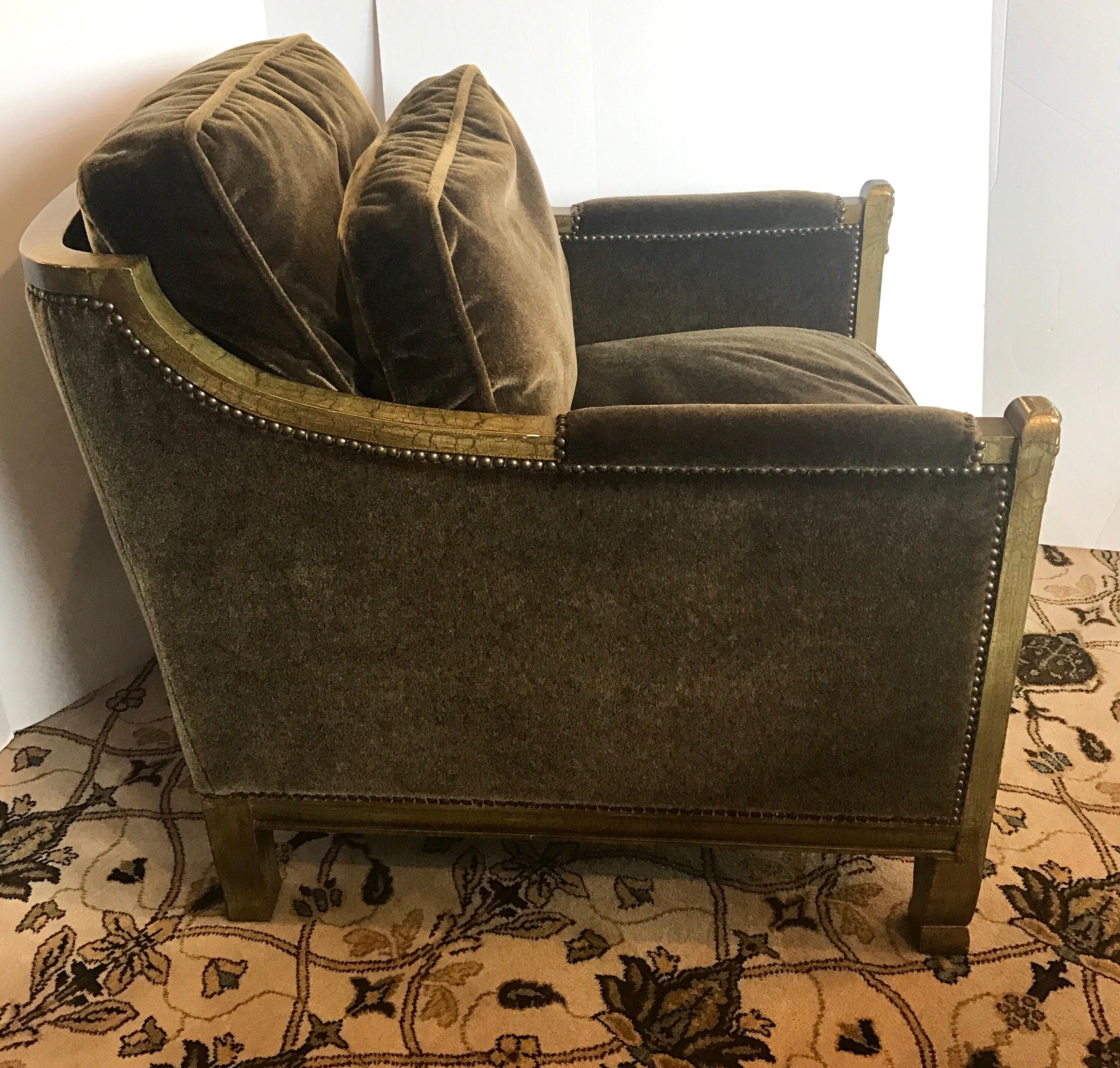 Pair of chic neoclassical style club chairs with a carved wood frame in a distressed gold finish and upholstered in a luxurious mohair velvet in an olive green color. Trimmed in brass nail heads all around. Very comfortable with three loose down