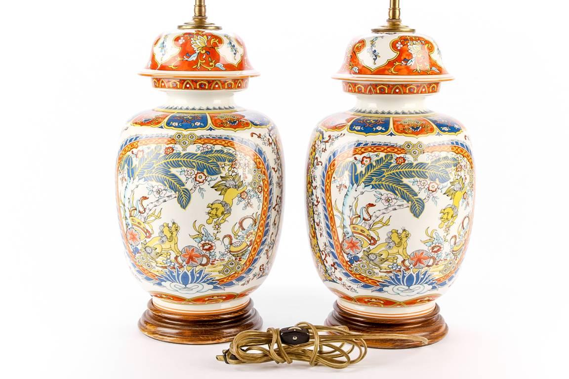 Pair of oriental porcelain ginger jars mounted as lamps, twin-light lamps, the jars decorated with foo lions facing off in a landscape with trees and flowers, in a bright blue and orange palette, lids and shoulders with floral panels, mounted on