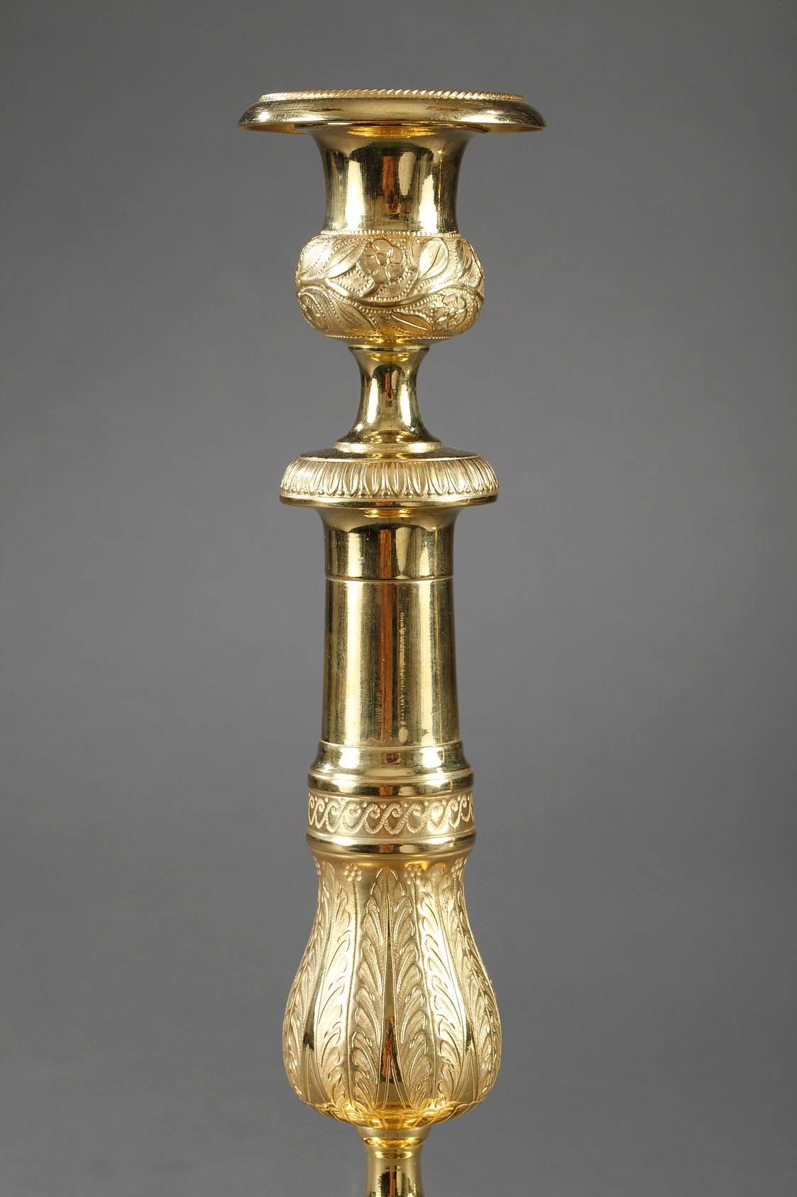 Coppia di candelieri in bronzo dorato e scolpito. Il fusto è ornato da un fregio di onde greche che separano la parte superiore, lucida, da quella inferiore, riccamente decorata con palmette, fiorellini e gadoning. È sormontato da una presa