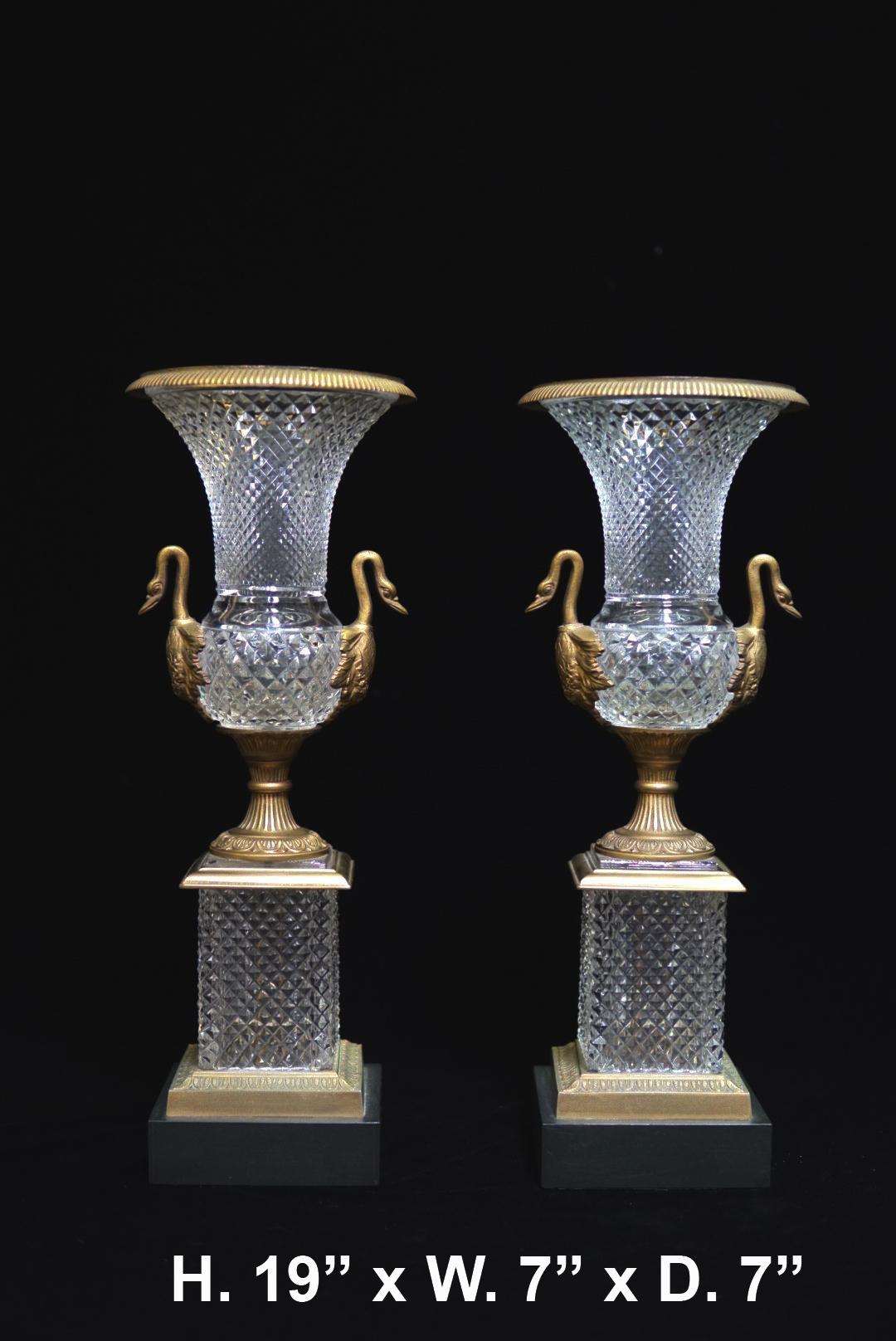 Impressive pair of French Louis XVI style ormolu mounted cut crystal urns with swan handles. Each urn is surmounted with a reeded gilt bronze rim over a cut crystal body flanked by two swans heads, above a square base and raised on bronze base.