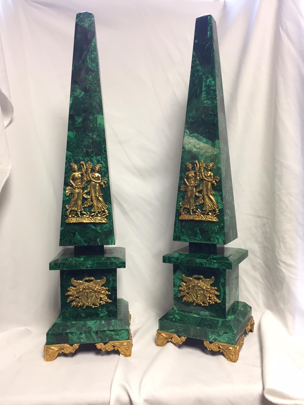 Imposing pair of ormolu-mounted malachite veneered obelisks. Top quality malachite veneered points centred by ormolu-mounted plaques portraying two playful maidens in robes holding floral garlands and Horn, over stepped base mounted with ormolu