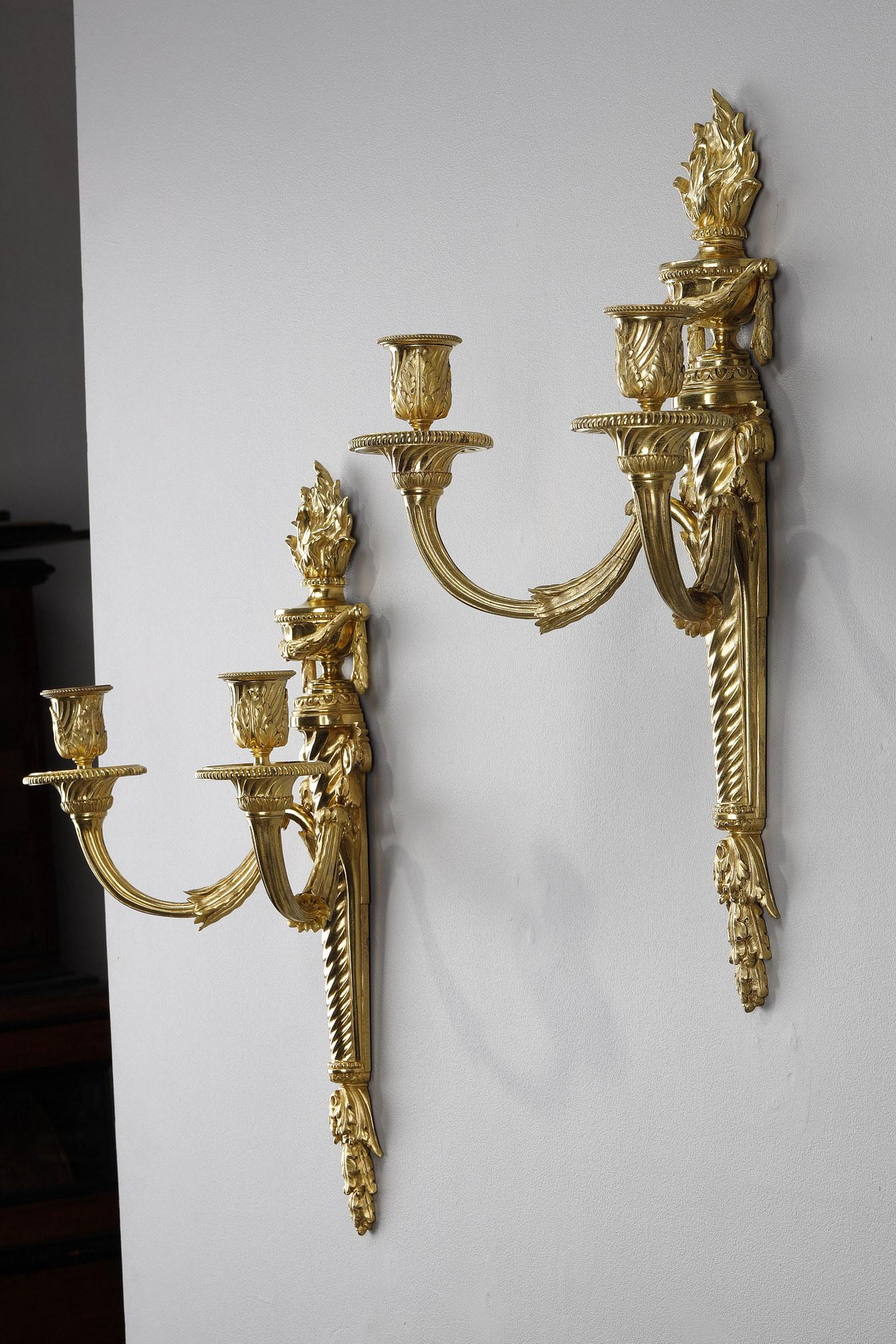A pair of ormolu and chased sconces with two light arms. The twisted plate is decorated with foliage and volutes. It is surmounted by a fire pot decorated with garlands of laurels. The two fluted light arms end with pearl motif bobeches and acanthus