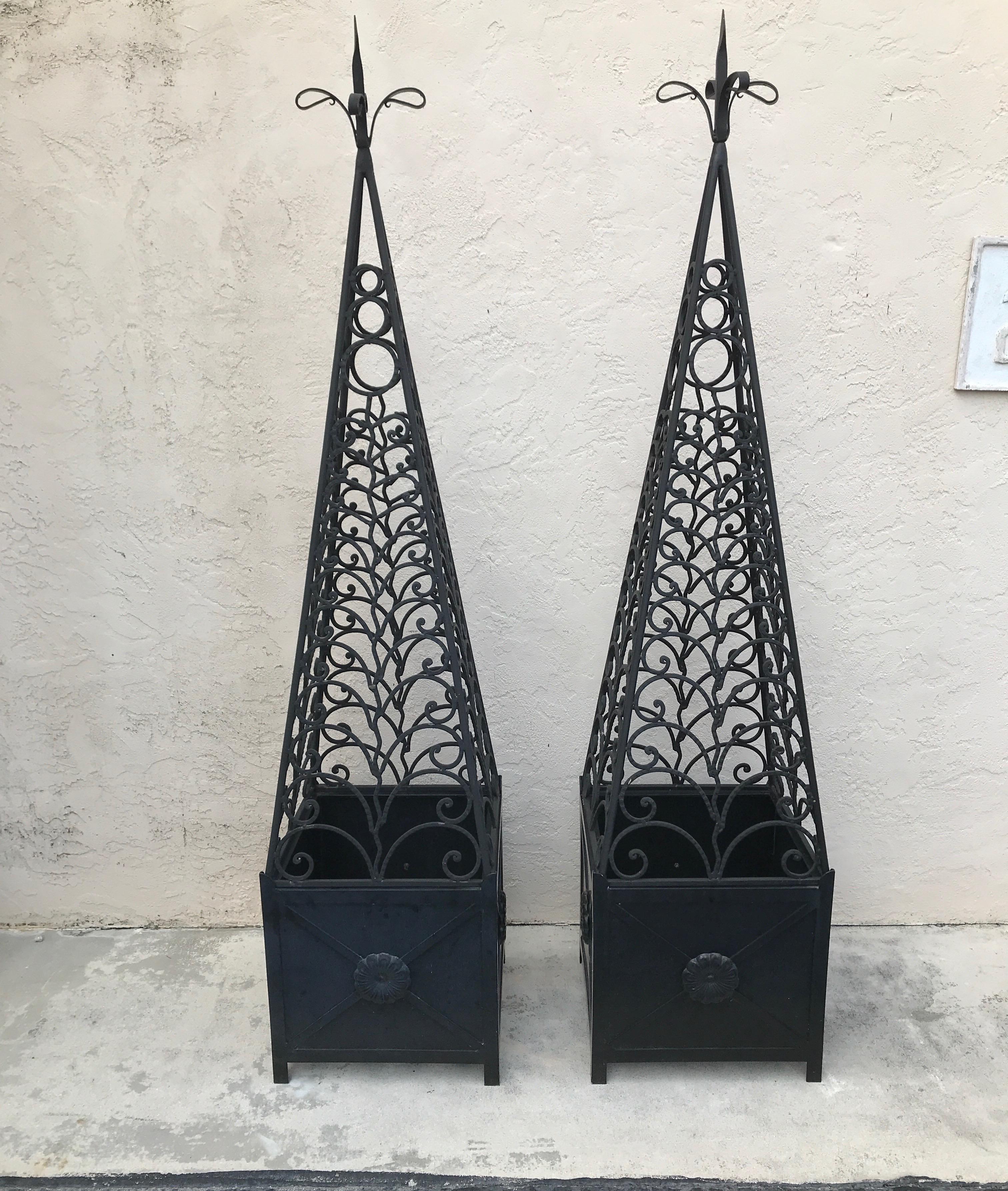 Imposing pair of black wrought iron obelisks sitting on square aluminum and iron planter bottoms.