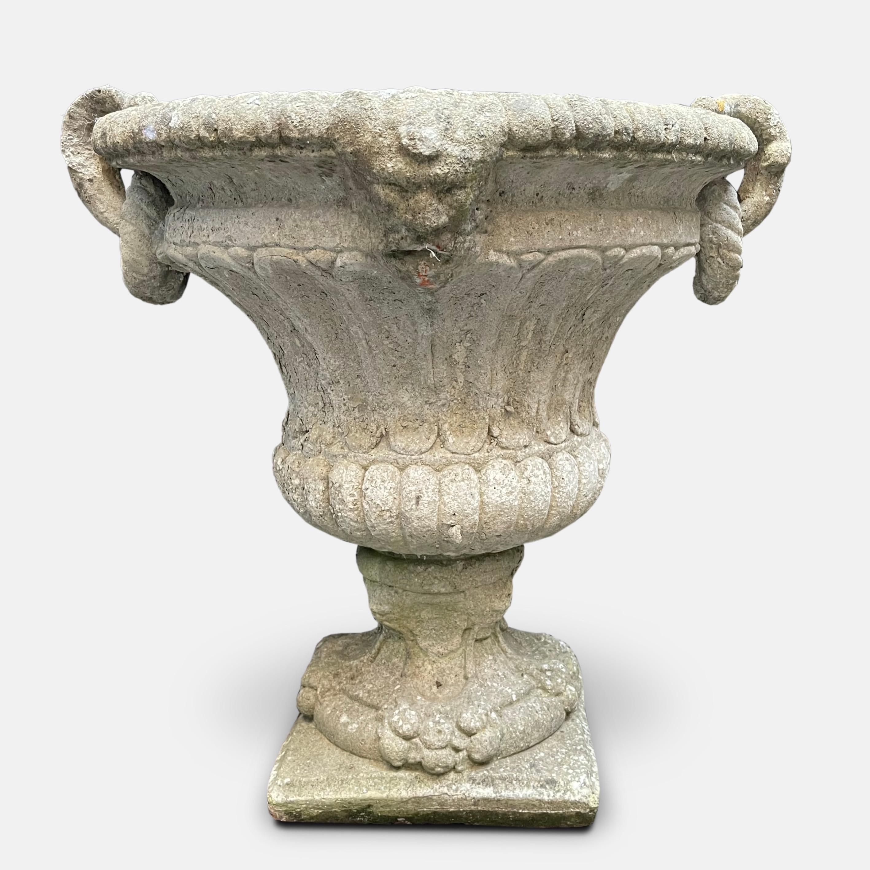 A pair of ornate early 20th century urn planters, each on a pedestal base, with twin handles and a thick ring shaped like a loop of rope through each handle, and formed, like the body of the urns from moulded reconstituted stone.
 Dimensions: H 68cm