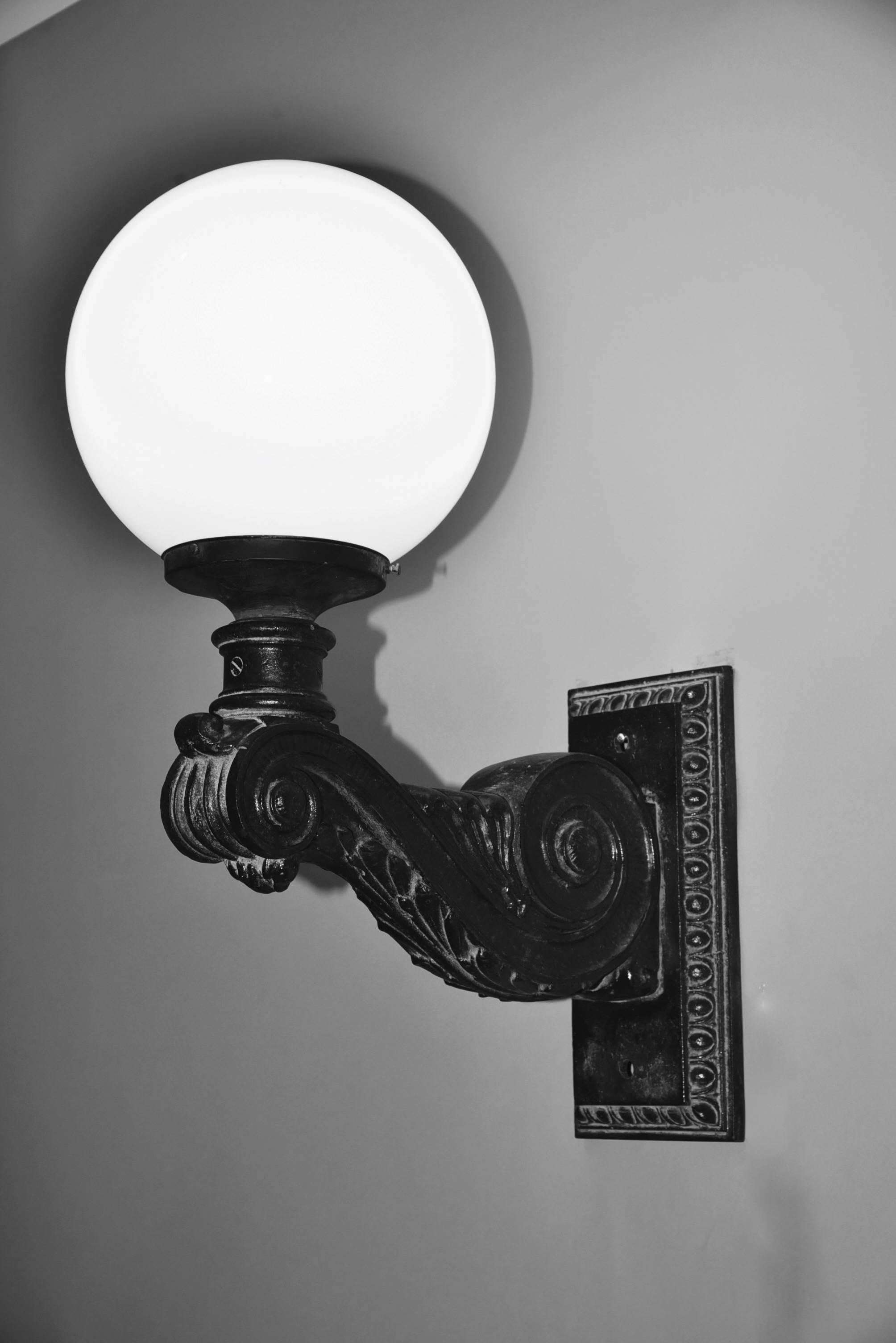 This is a rare pair of large heavy industrial black cast iron wall-mounted lamps or sconces for outdoors from the 1920's. Black cast iron rectangular egg-and dart bordered back plate issuing scrolled acanthus leaf detail arm supporting a globe opal