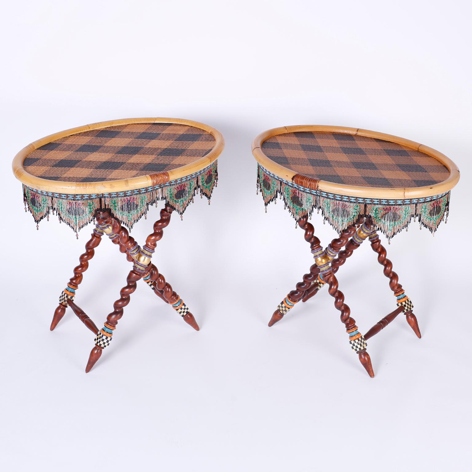 Pair of whimsical collapsible serving tray top or end tables with argyle wicker surfaces bordered with bamboo and glass bead fringe. The carved wood stands have barley twist legs with unexpected painted decorations. Branded Mackenzie-Childs on the