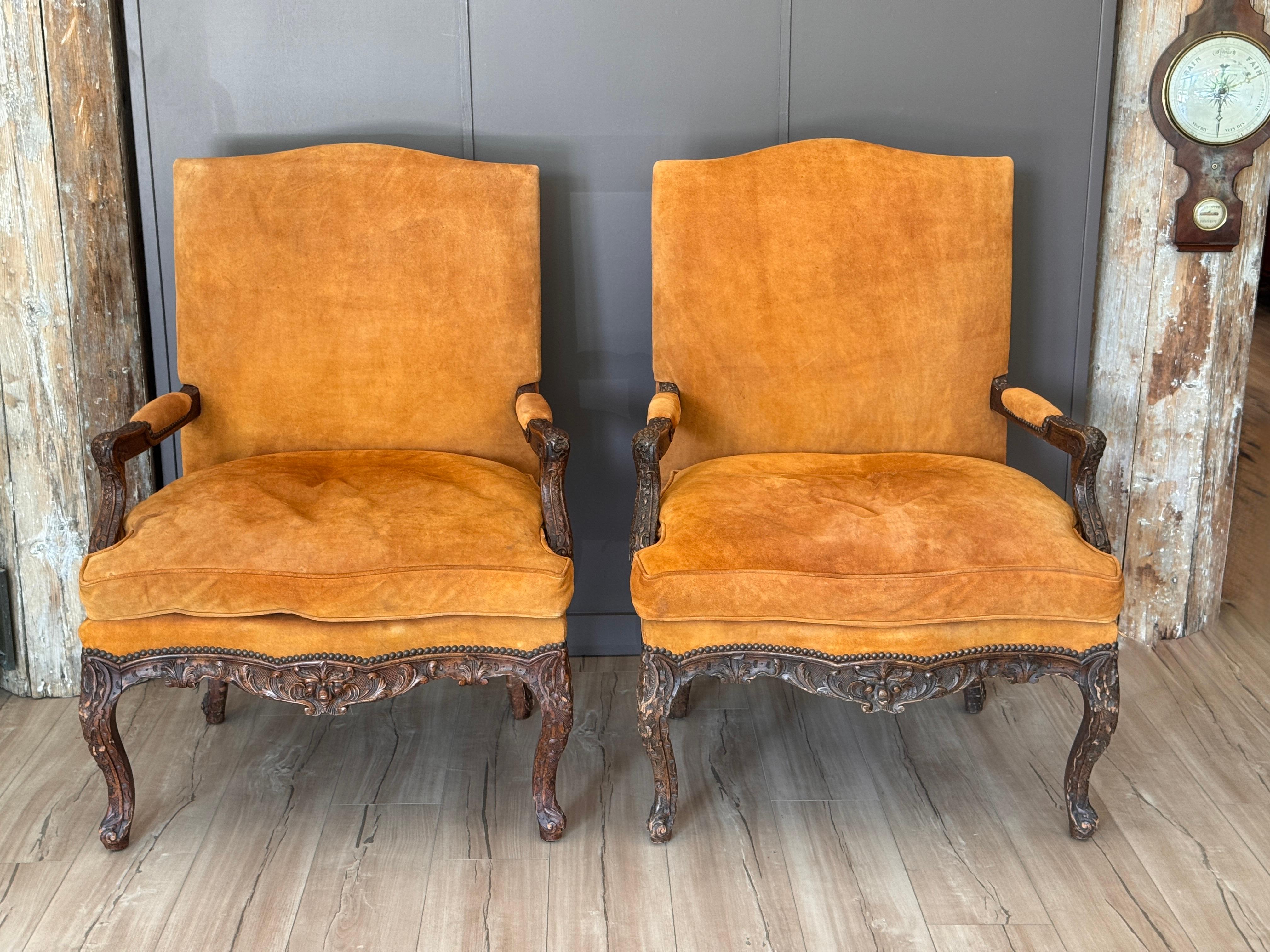 A generously scaled pair of 19th century French Louis XV style fauteuils featuring boldly carved cabriole legs, scroll arms, and a detailed foliate skirt with floral and shell motifs. The frames are upholstered in a soft, deep orange leather fabric