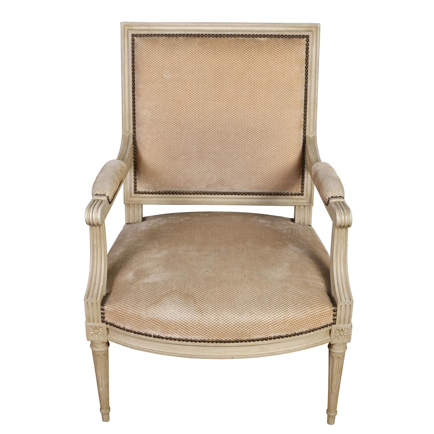 A pair of oversized, painted French arm chairs circa 1940. The pair of Louis XVI chairs are painted off white, with square backs, reeded and carved frame, arms and tapered legs. Each leg meets the seat frame with a carved floral motif. Arm rests are