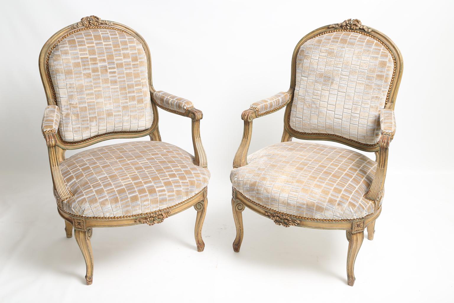 Pair of armchairs, having a painted finish showing natural wear, each arched, channelled frame surmounted by carved ribbons, padded backrest flanked by scrolling arms with padded elbow rests, raised on fluted, down swept terminals, its stuff-over