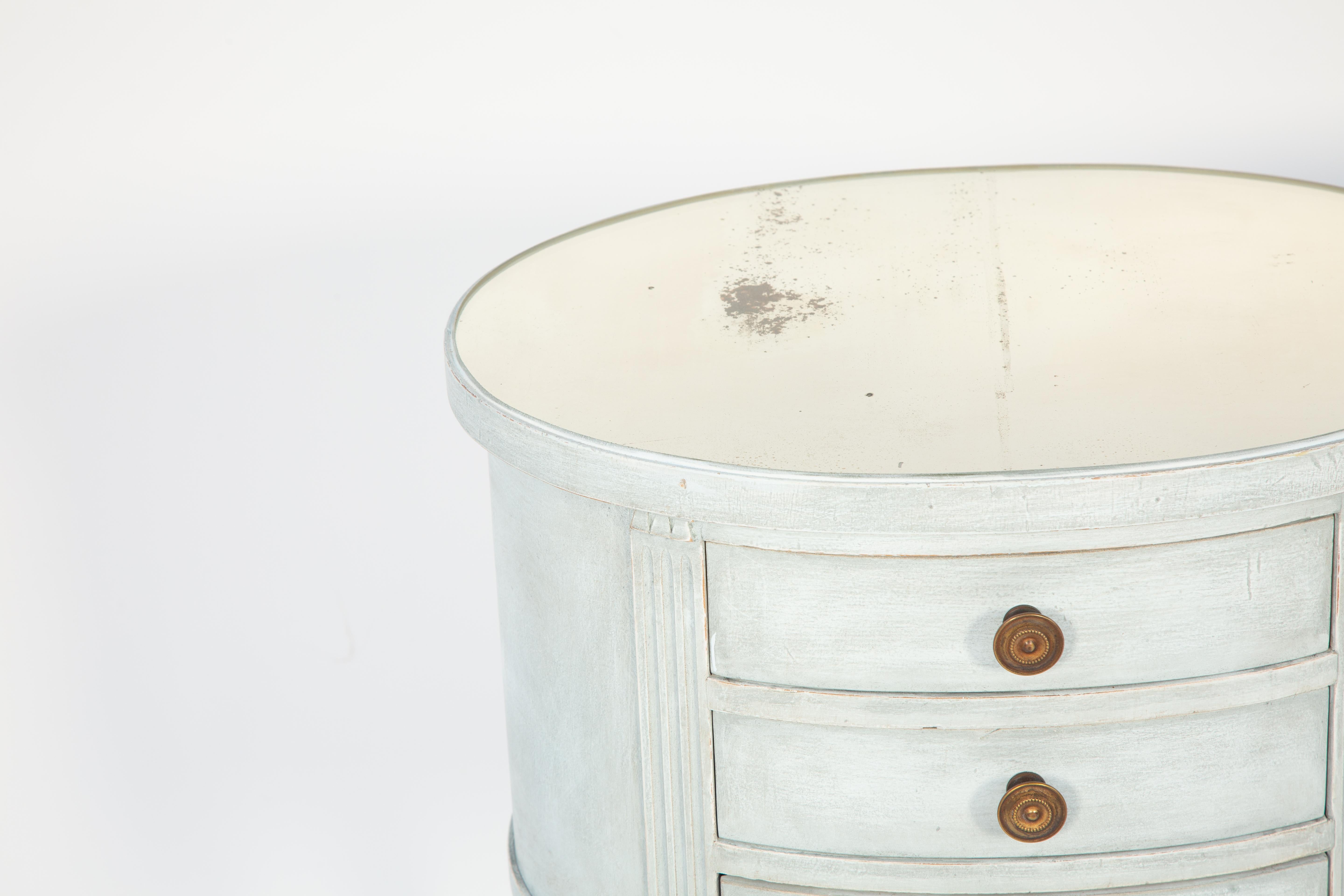 Pair of vintage bedside tables, painted pale blue finish showing natural wear; each having an oval top of antiqued mirror, on conforming table base with triple-stacked drawers flanked by fluted stiles, raised on round, tapering, fluted legs joined