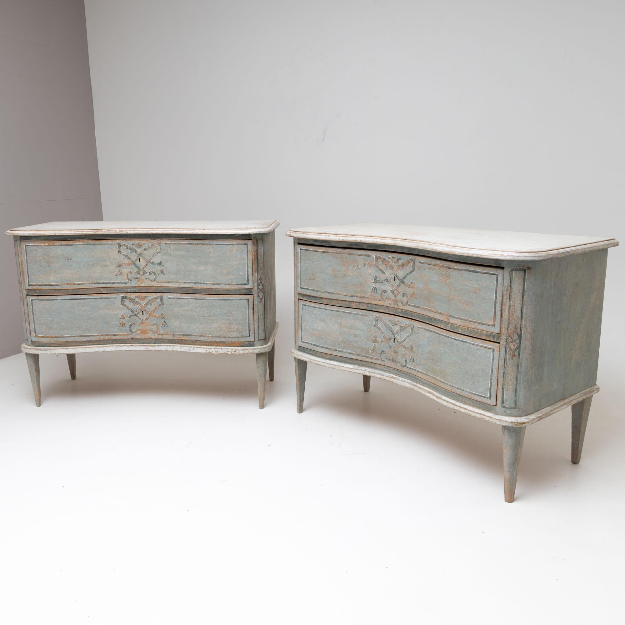 Pair of chests of drawers with two drawers and curved fronts. The chests of drawers rest on square tapered feet and have been repainted and given an antique patina. The light blue setting is contrasted by white accents. The decors are based on
