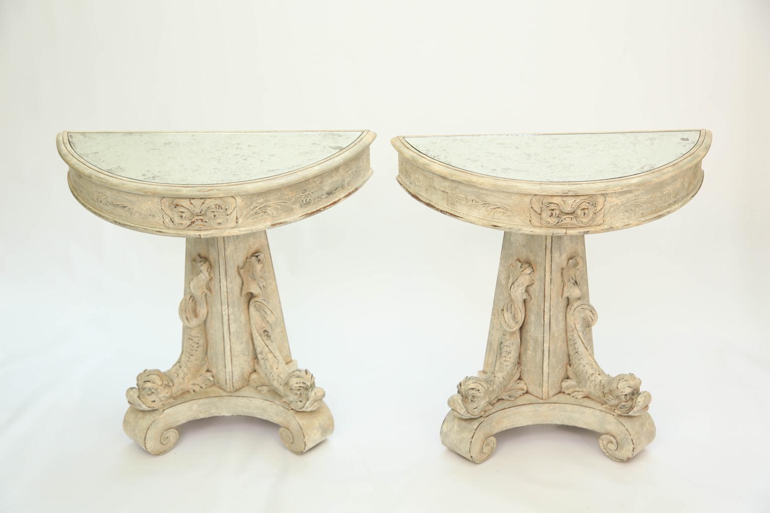 Pair of demilune consoles, with painted finish showing desirable wear; each having a D-shaped top of aged mirror, conforming foliate-carved apron, raised on pedestal base, decoratively carved with classical dolphins, upon a concave base ending in