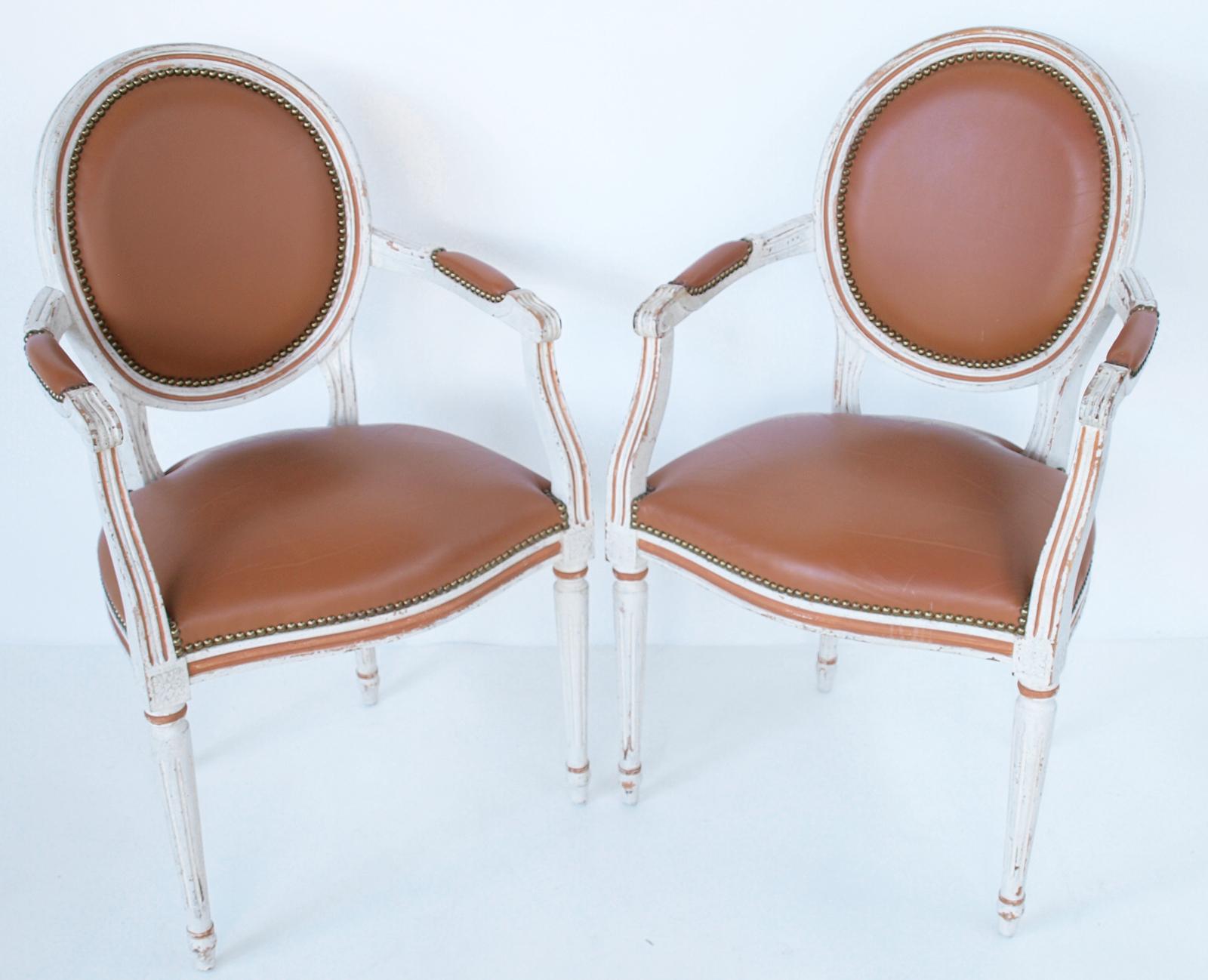 Pair of armchairs, having a painted finish showing natural wear, upholstered in leather with nailheads, each channeled frame inset by an oval padded backrest, oustwept arms with elbow cushions, finished in scrolls, supported by S-scroll terminals