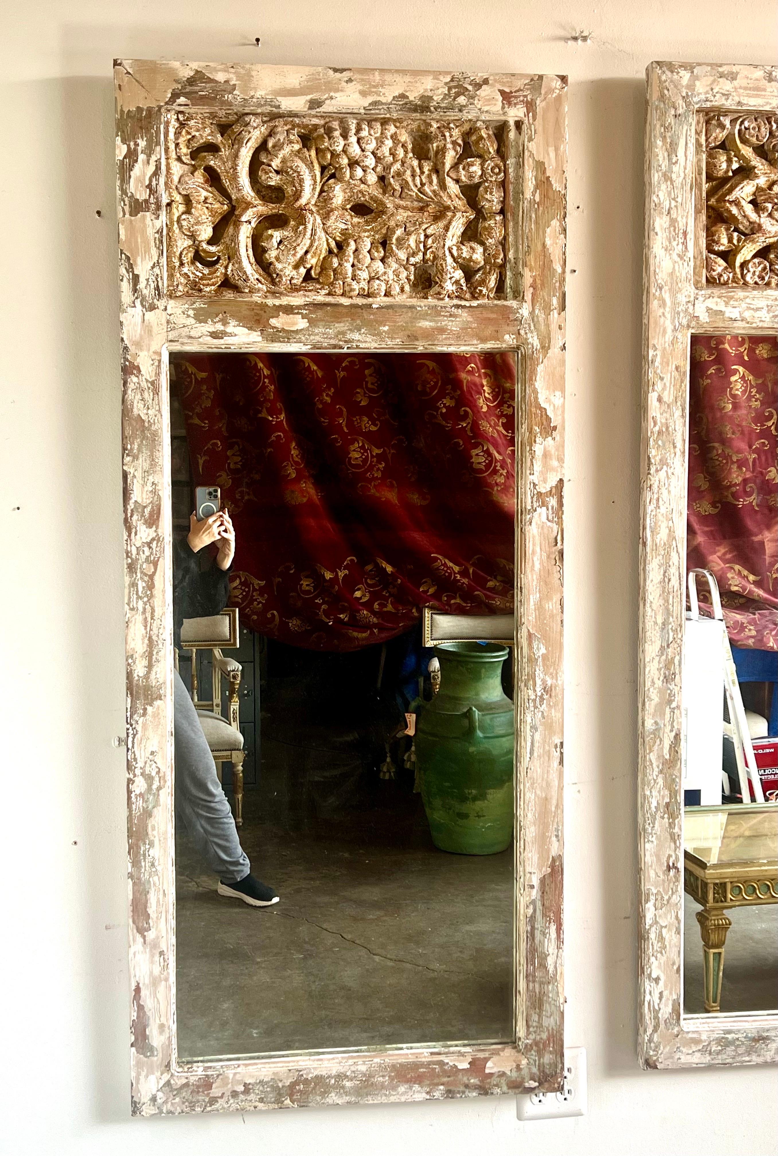 Pair of tall, rectangular mirrors, each adorned with ornate, gilded carvings at the top.  The carvings feature classical motifs, including acanthus leaves and floral design, which add a touch of opulence and historical elegance.  The painted frames