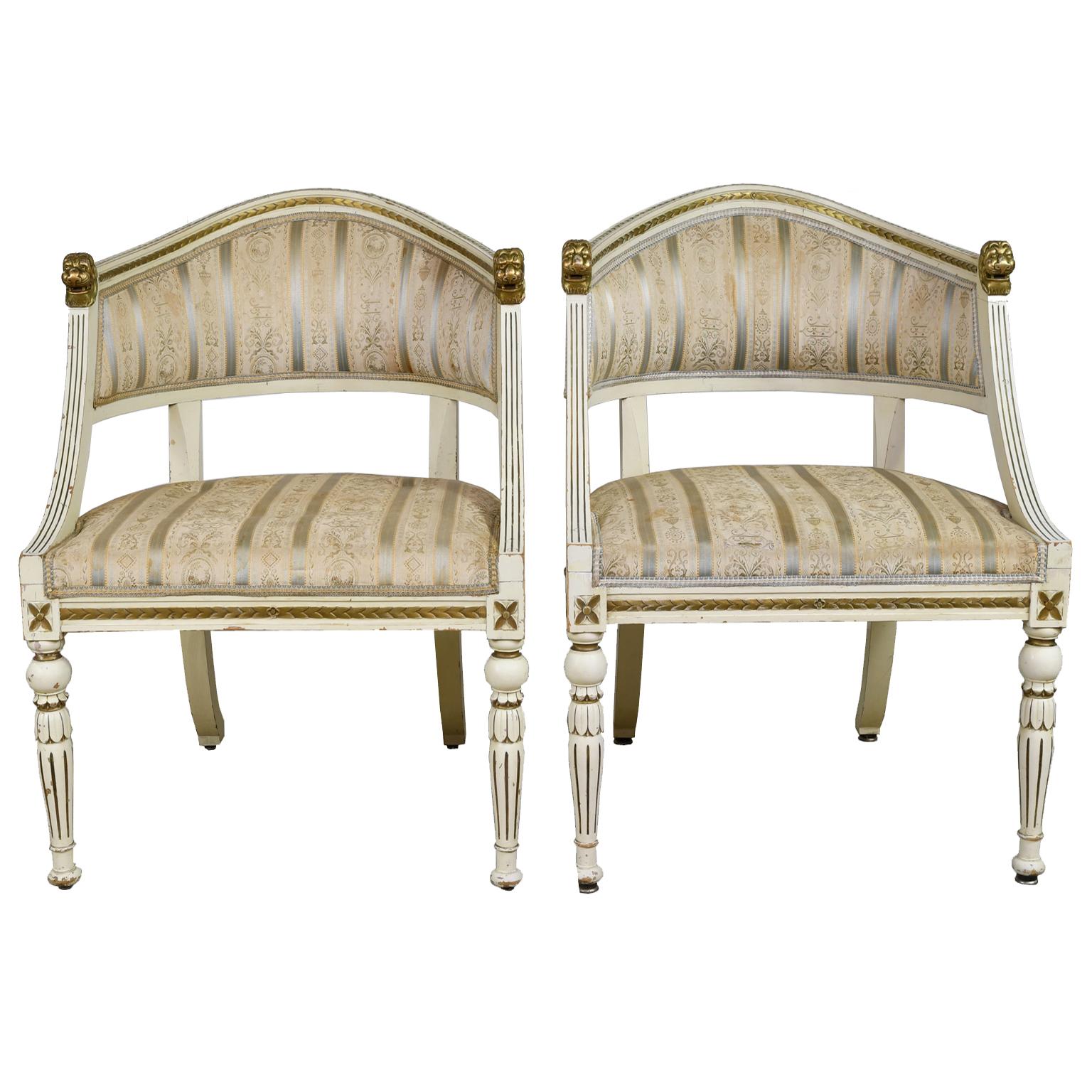 A very handsome pair of Gustavian-style armchairs with upholstered seat and back. Frame is painted an ivory color with antique gold accents and features a gondola form with arched back, carved lions heads at each end of the crest rail which has a