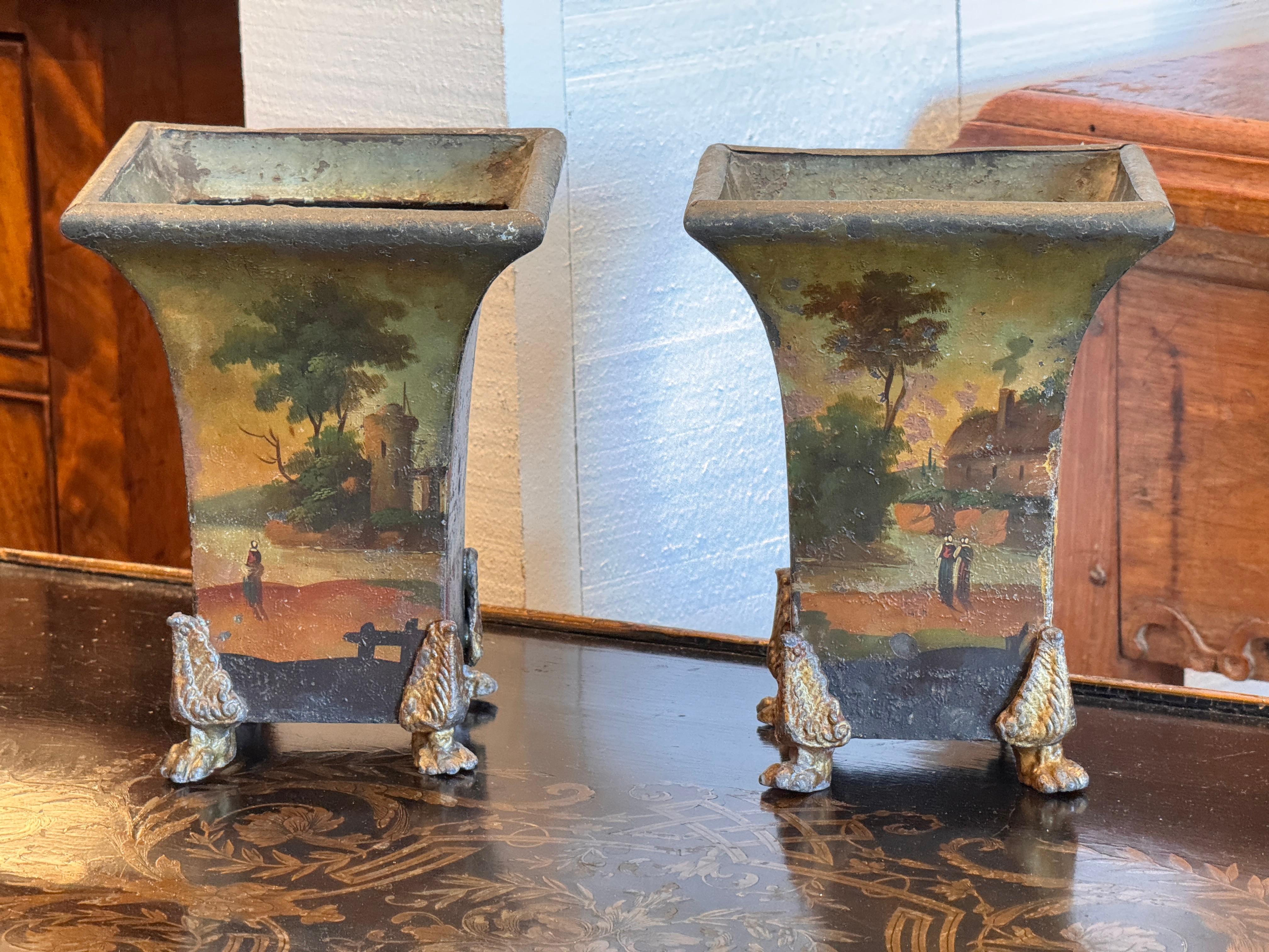 Decorativa pareja de urnas de tole pintadas a mano, cada una con paisajes románticos en todas sus caras. Se eleva sobre unas características patas doradas. La superficie presenta un atractivo desgaste propio de la edad, que realza el carácter de las
