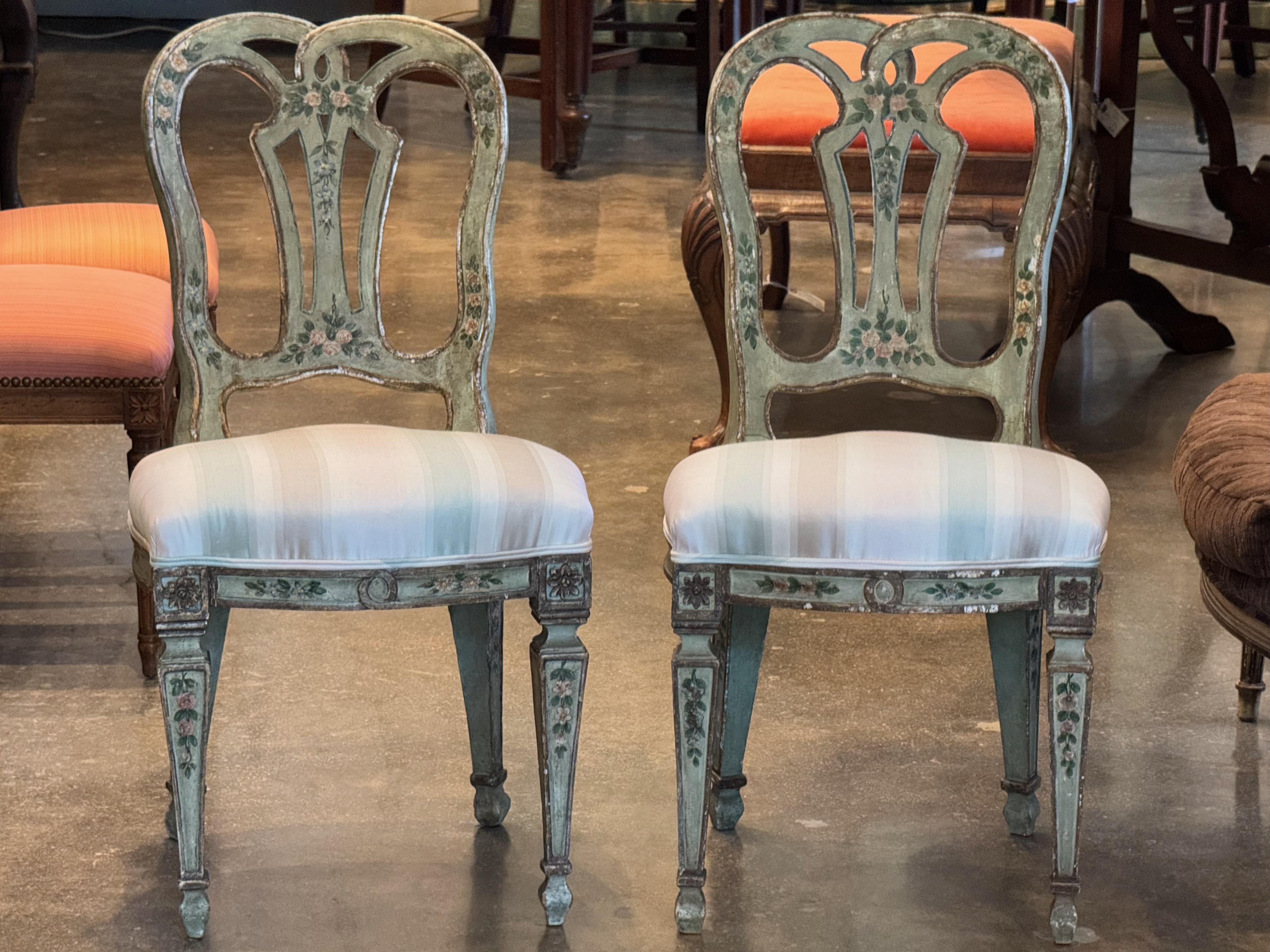 Paire de chaises d'appoint vénitiennes décoratives avec dossier ouvert sculpté et motifs floraux peints à la main. La peinture verte usée ajoute du charme et de la patine, tandis que les sièges sont recouverts d'un tissu à rayures pâles pour un
