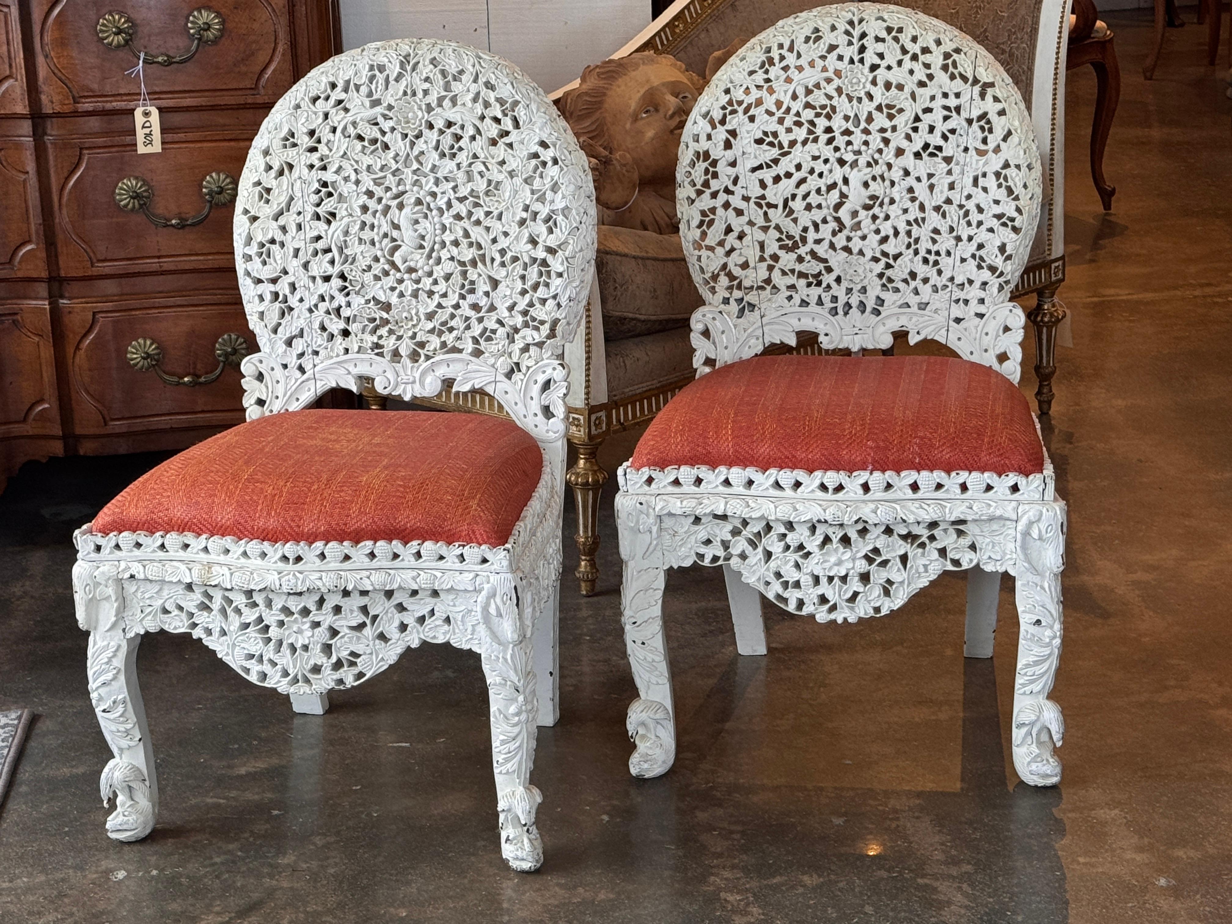 Pair of painted white carved side chairs with densely pierced floral work across the backs, aprons, and legs. The carving includes layered foliate patterns and scroll details, with front legs ending in carved paw-like feet. Close-up images show