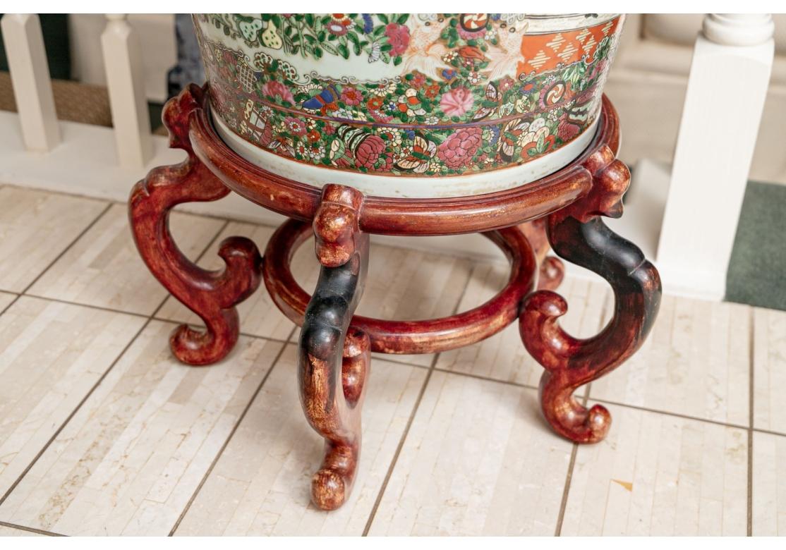 An extraordinary pair of oversized Rose Mandarin Vases with several scenes of Asian figures and landscapes in bright and colorful designs. Bold figural dragons and guardian lions to the neck of the vase, scallops to the rim. Rests on a footed wood