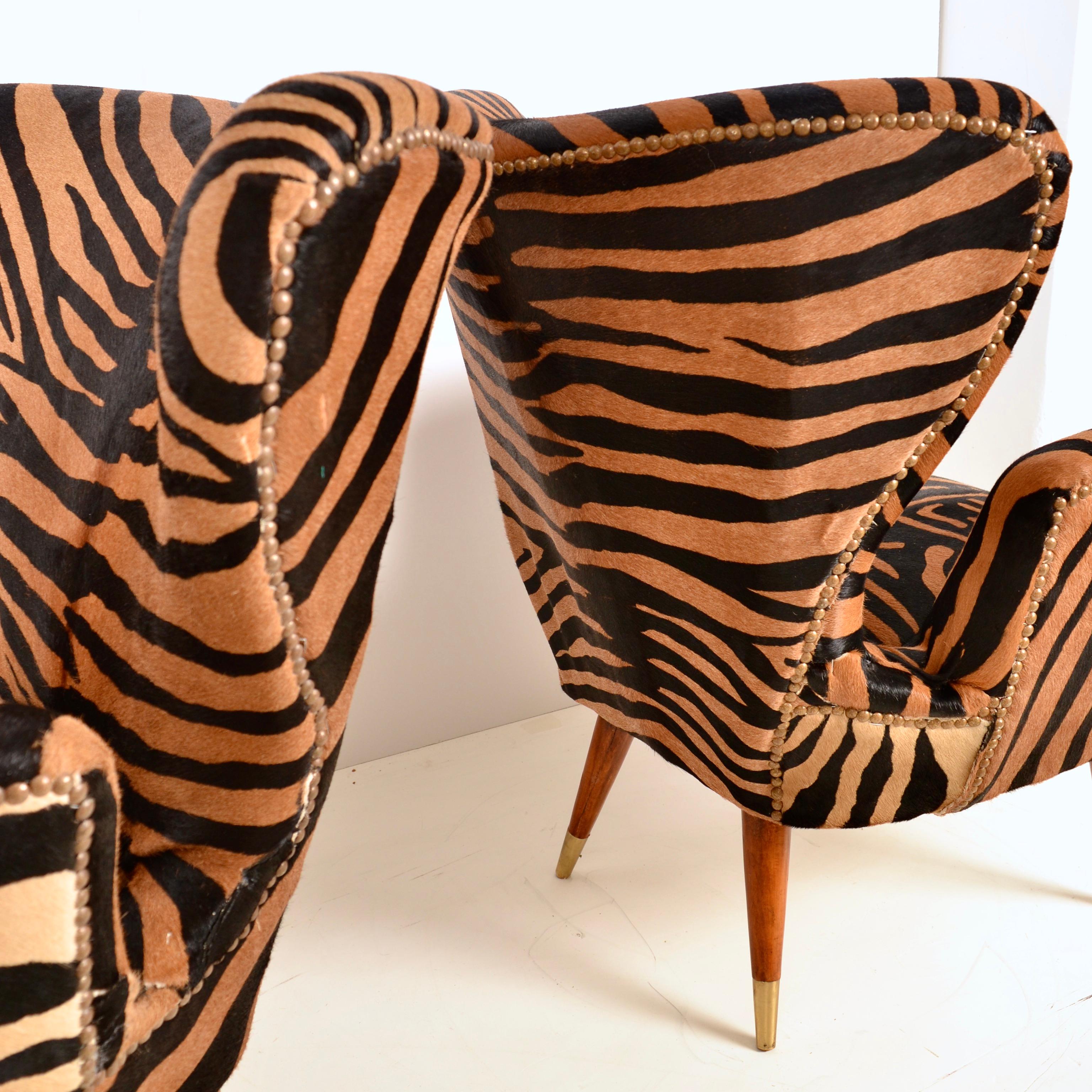 This sculptural pair of winged arm chairs were created by the influential Italian designer, Paolo Buffa. Newly upholstered in zebra patterned, dyed cow skin with nail head details, the chairs feature tapered wood legs with brass caps. Iconic design