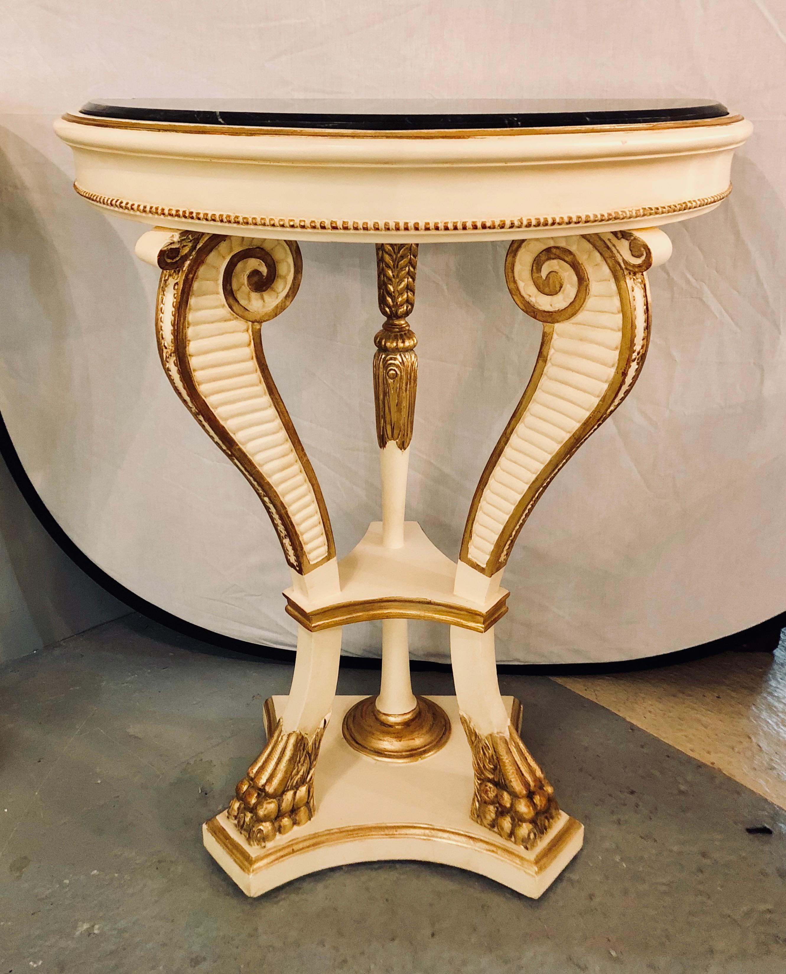 A pair of parcel paint and gilt decorated marble top demilune console tables with claw feet. Each having a linen white painted background with a gilt gold design having a black marble top in the demilune form. Supported by scroll reeded legs.