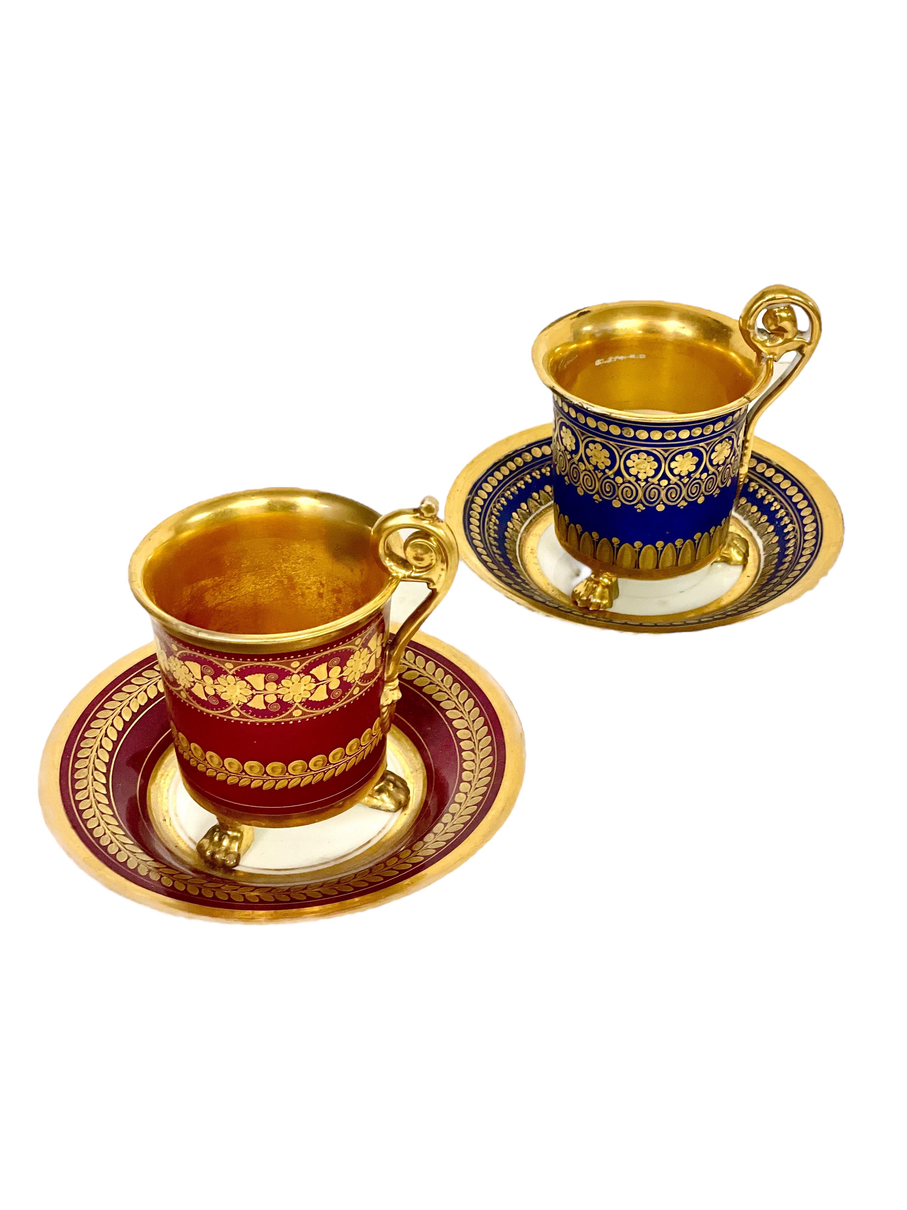 A superb pair of opulent 'Porcelain de Paris' three-footed coffee cups with matching saucers – one blue, one burgundy – each richly embellished with hand-painted gilt detailing in eye- catching geometric bands. These very stylish cups are also fully