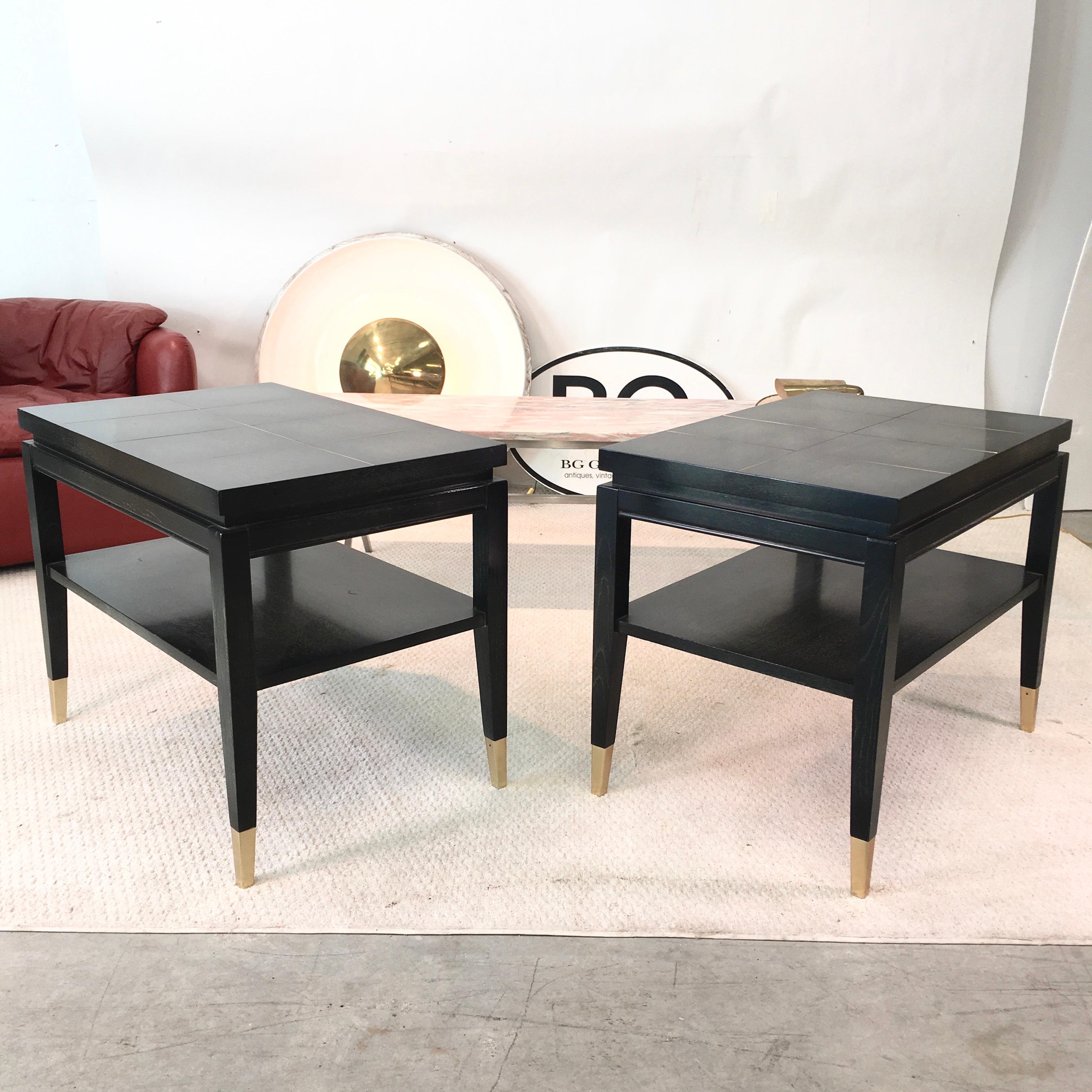 Pair of 1950s well-proportioned rectangular two-tier side or end tables in the style of Tommi Parzinger, stained black with transparent finish highlighting the grained wood with brass tone metal inlay and sabots on four square tapered legs. Lower